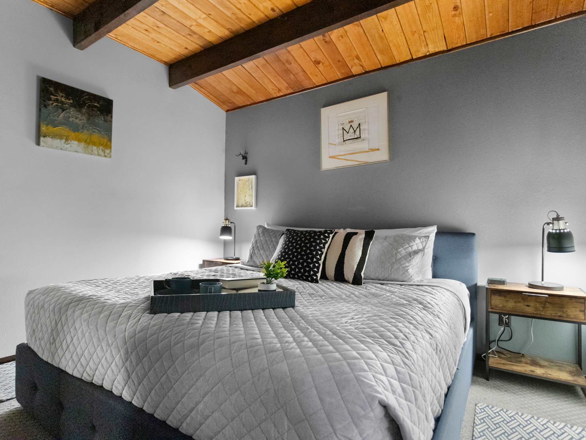 A cozy bedroom features a light blue upholstered bed with a quilted grey comforter and decorative pillows, flanked by dark nightstands with modern lamps. The room has a grey accent wall, exposed wooden ceiling beams, and framed artwork.