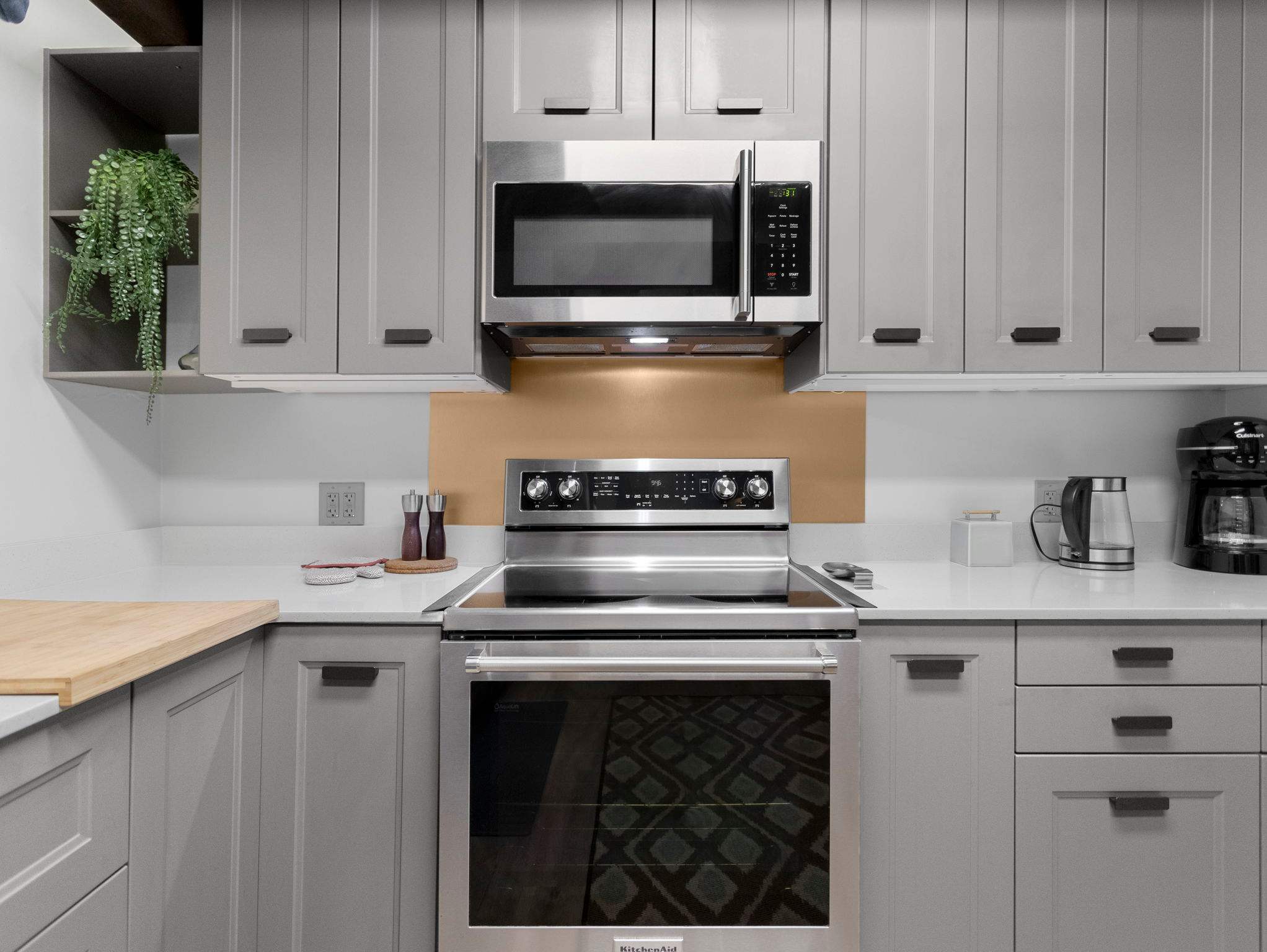 A close-up of the kitchen's cooking area, featuring a stainless steel electric range with a matching microwave oven above it. Light grey shaker cabinets flank the appliances, with a warm bronze backsplash and white countertops.