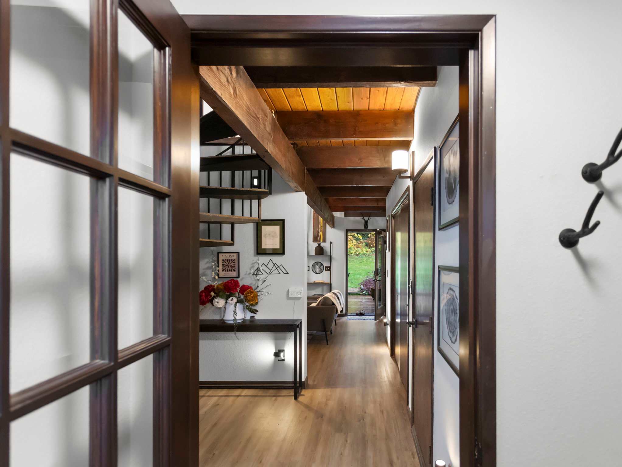 A hallway with hardwood floors and exposed wooden beams on the ceiling, leading towards a spiral staircase and a console table with decor. The view extends into another living area.