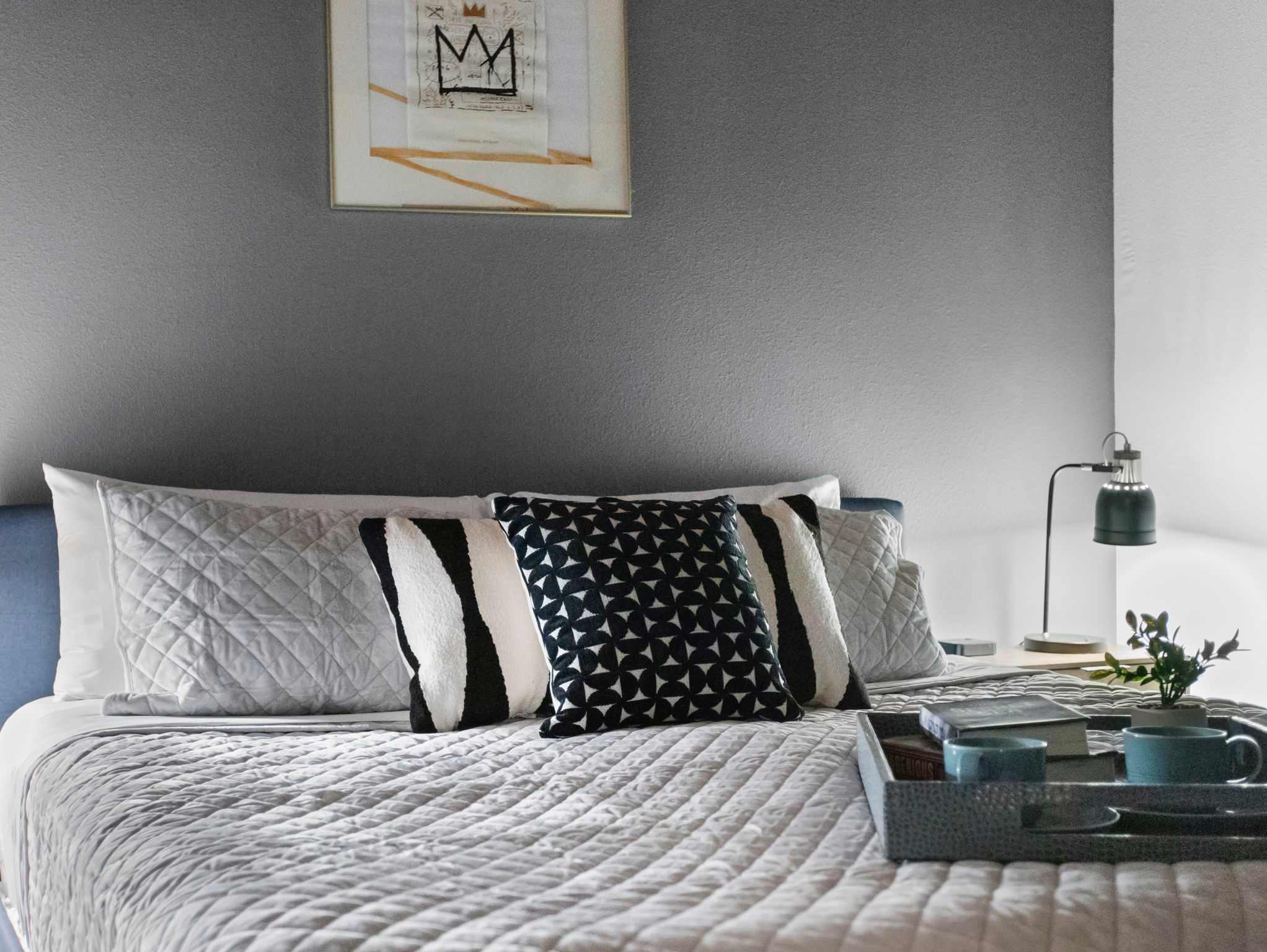 A bedroom with a large bed covered in a quilted white comforter and various patterned pillows, set against a gray accent wall. A bedside table with a lamp and a serving tray with mugs are visible.