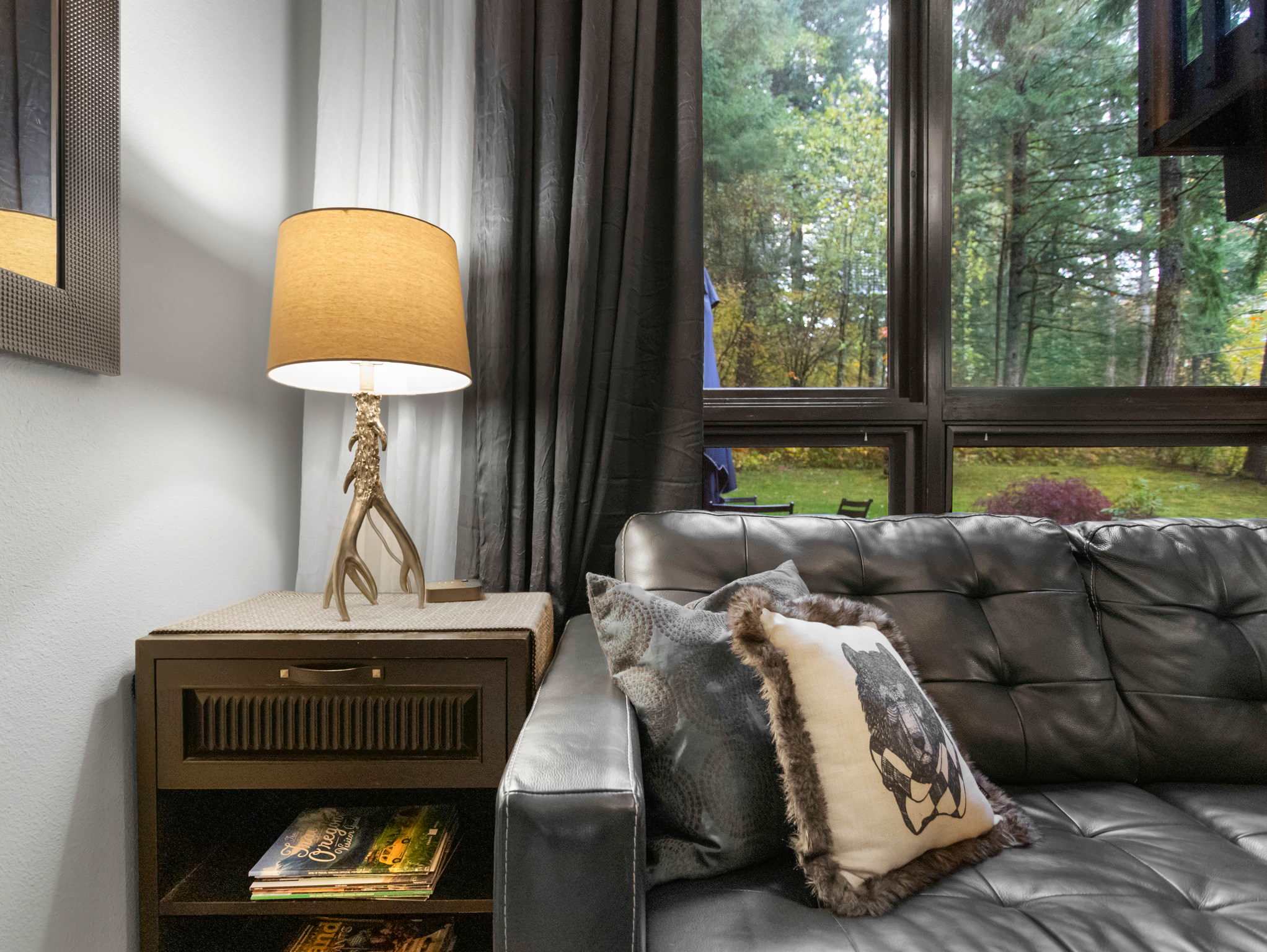 Detail shot of a black leather sofa with decorative pillows, a dark wood side table, and a unique table lamp with an antler-like base. Magazines are stored on the lower shelf of the side table.