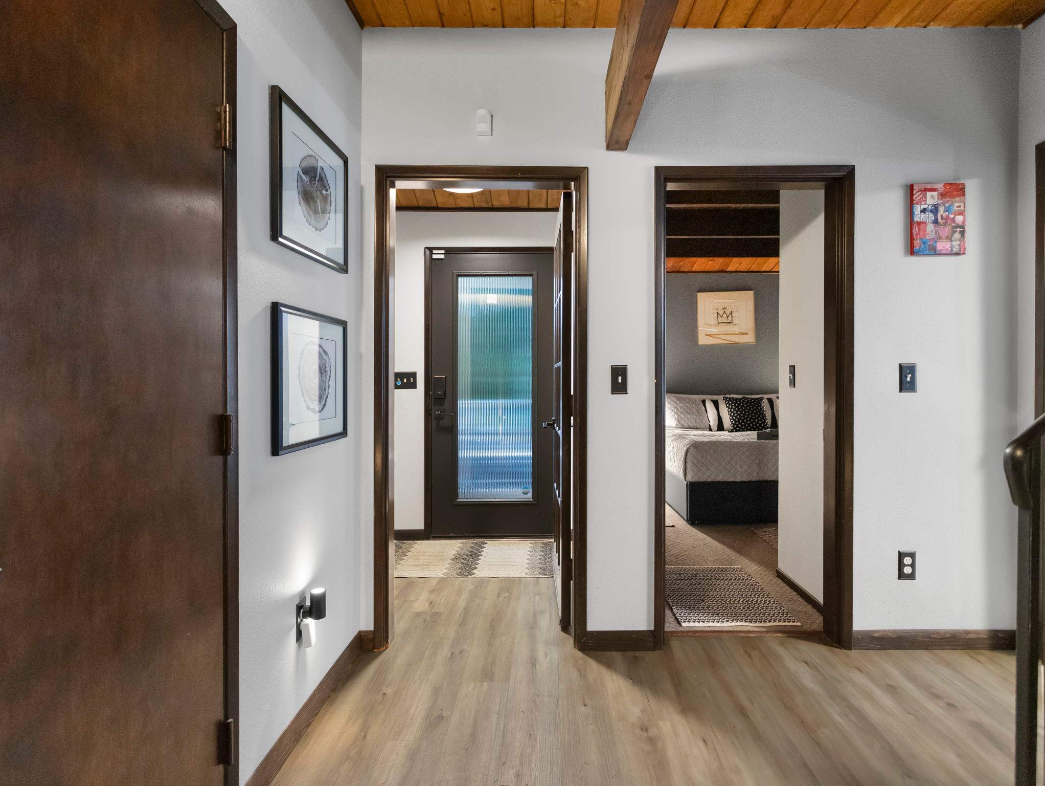 A bright hallway with light wood flooring and white walls leads to several dark-framed doorways, one opening to an exterior door. Exposed wooden beams are visible on the ceiling, and framed artwork decorates the walls.