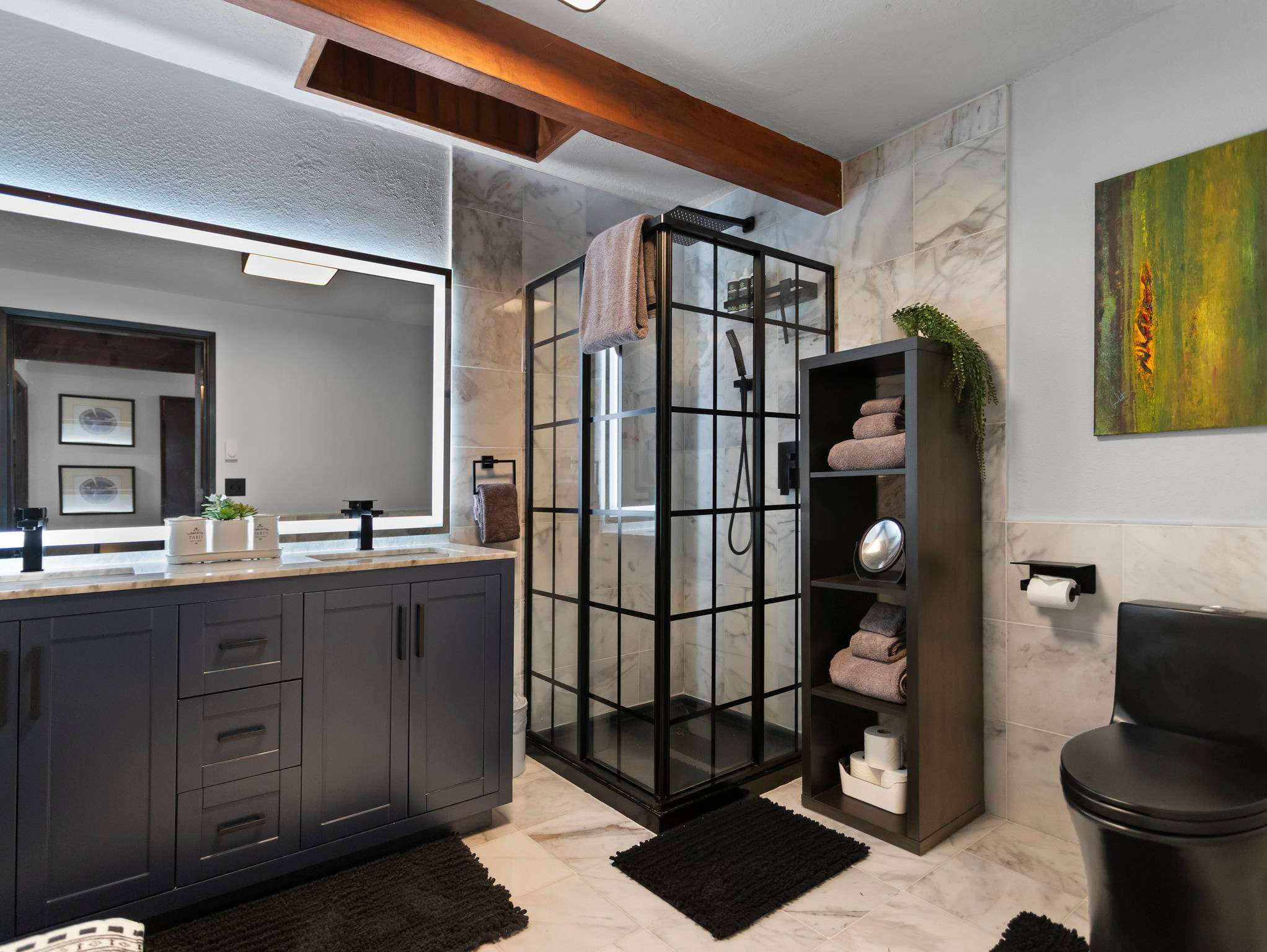 A stylish bathroom features a dark grey double vanity with a marble-look countertop and an illuminated mirror, alongside a black-framed glass shower enclosure. Black toilet, dark shelving unit with towels, and white marble-look floor tiles complete the modern design.