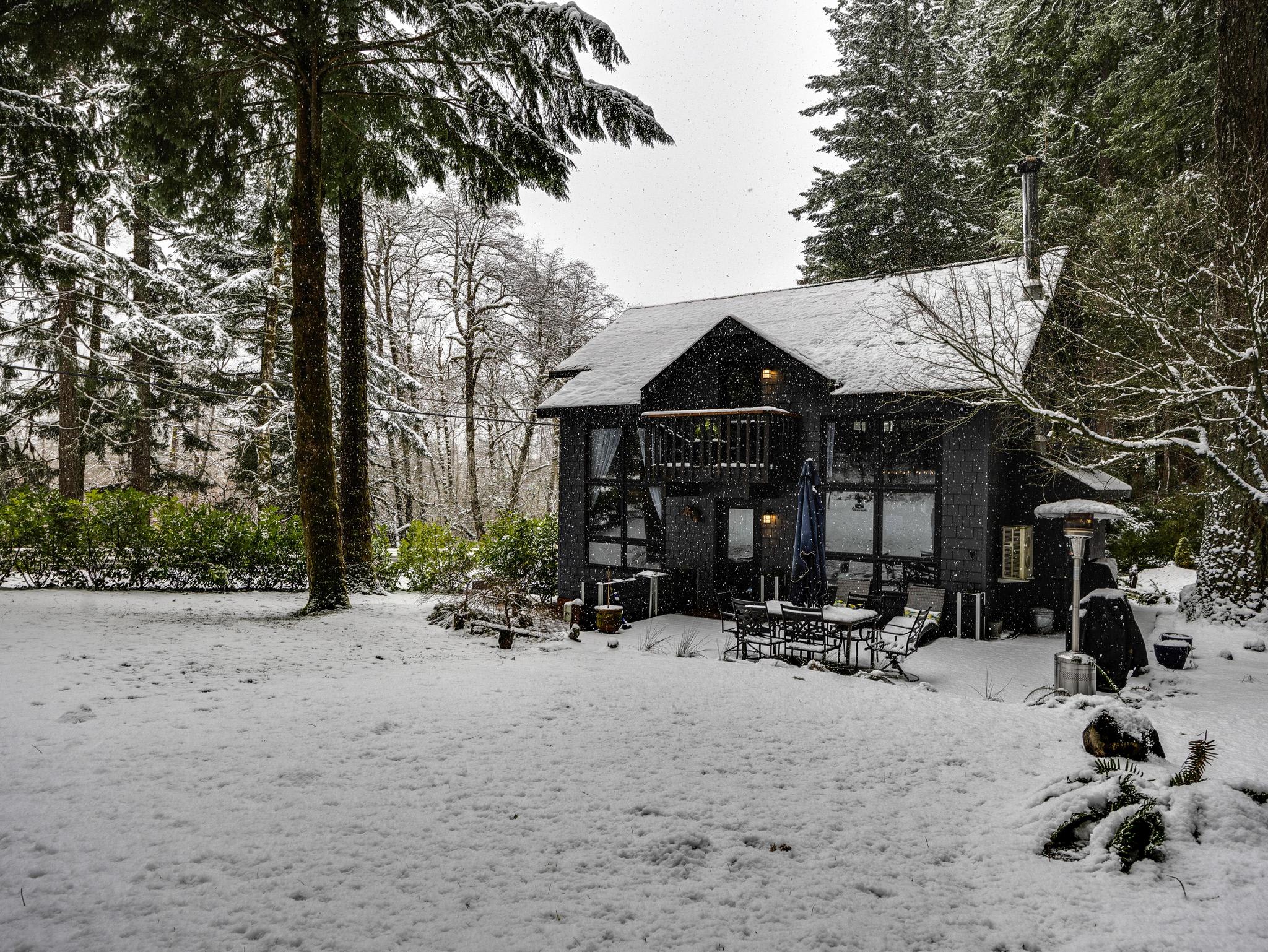 The dark gray house is covered in a fresh blanket of snow, with a snow-covered lawn and surrounding evergreen trees under a soft snowfall. A small balcony and outdoor seating are visible, creating a picturesque winter scene.
