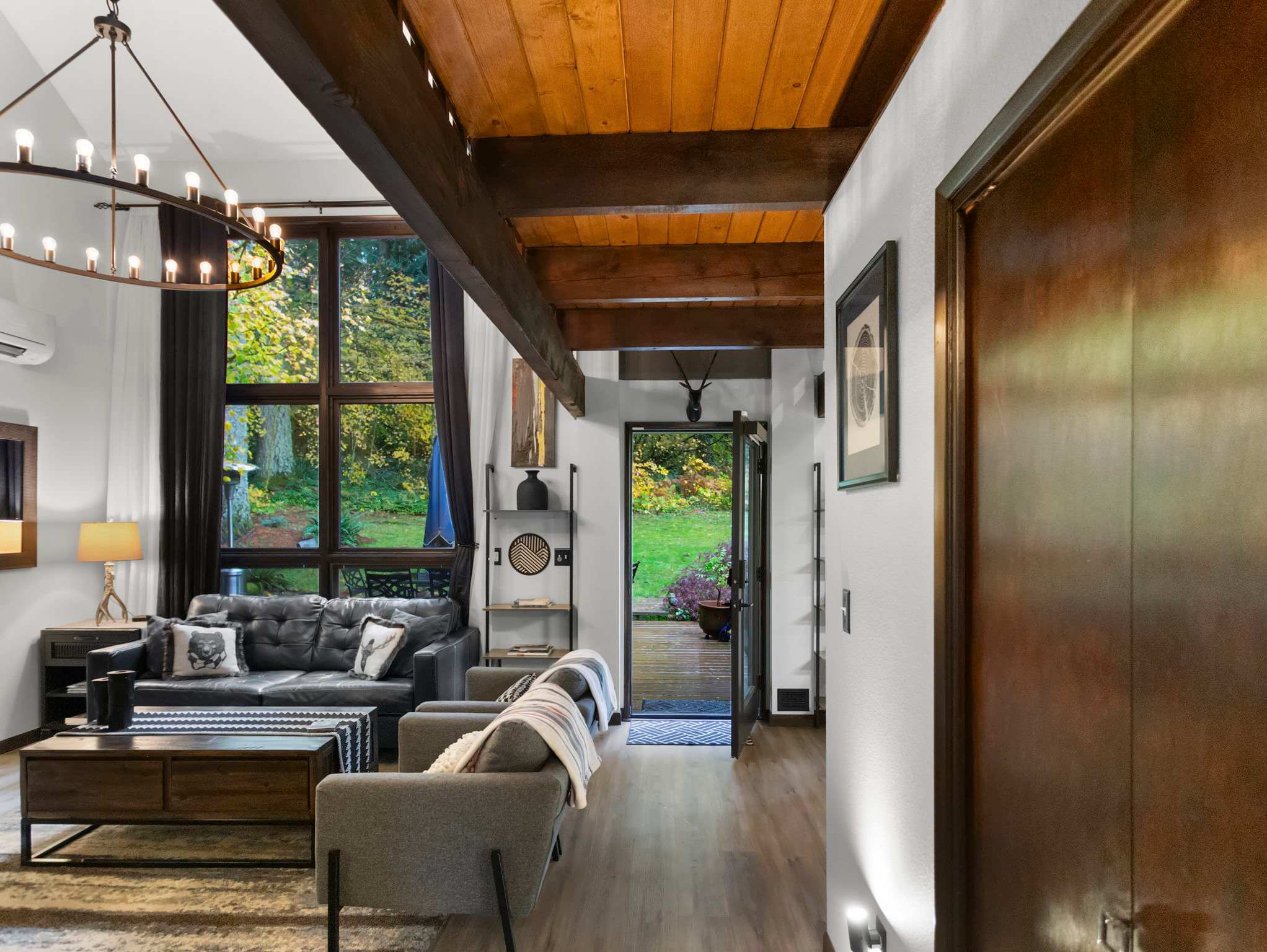 A bright living area with large windows overlooking the backyard, featuring a dark leather sofa and a gray armchair. Exposed wooden beams adorn the ceiling, and a doorway leads to the outdoor deck.