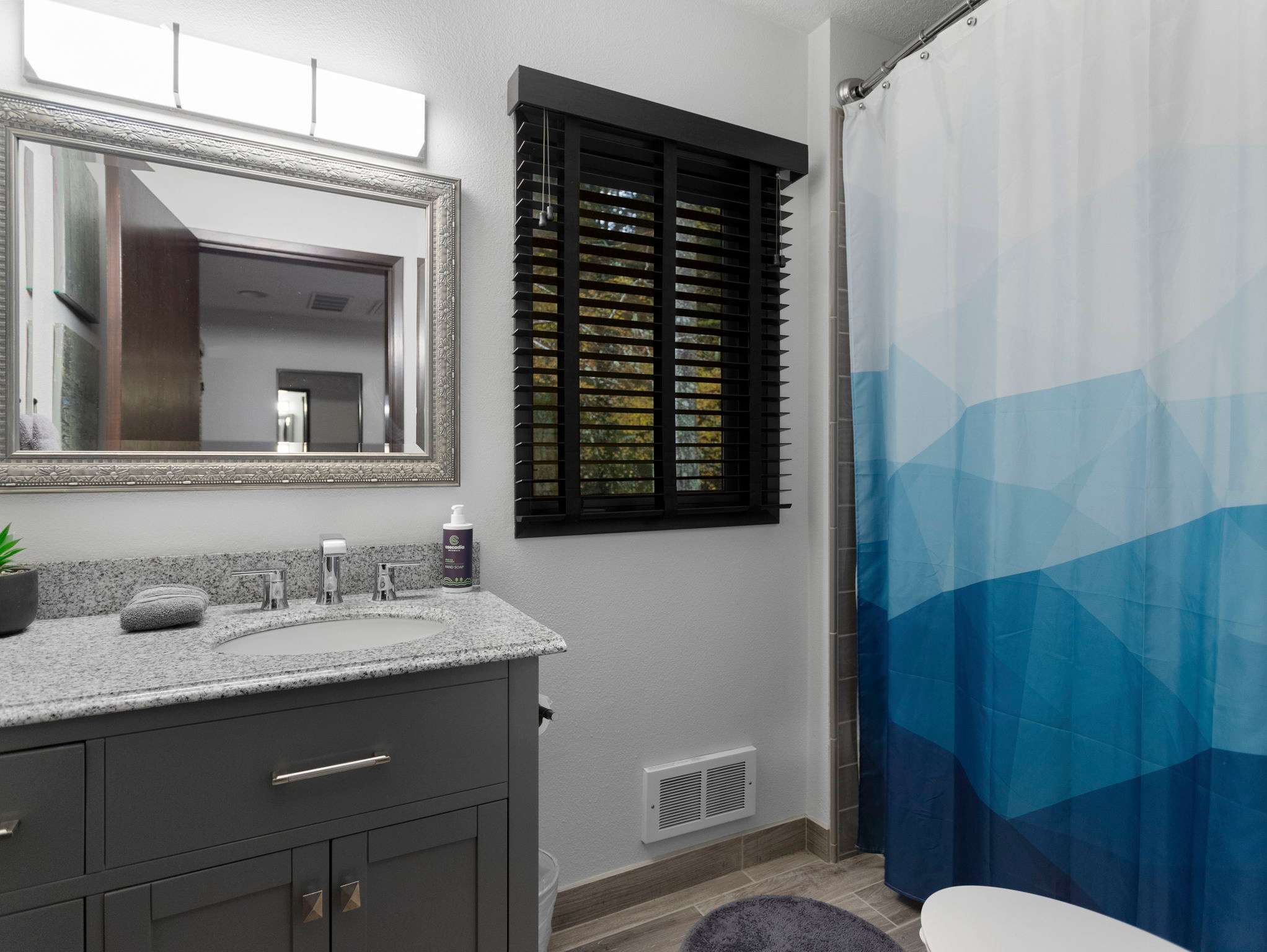 Modern bathroom featuring a gray vanity with a granite countertop, an oval sink, and a large silver-framed mirror. A window with dark wooden blinds and a blue and white geometric shower curtain are visible.
