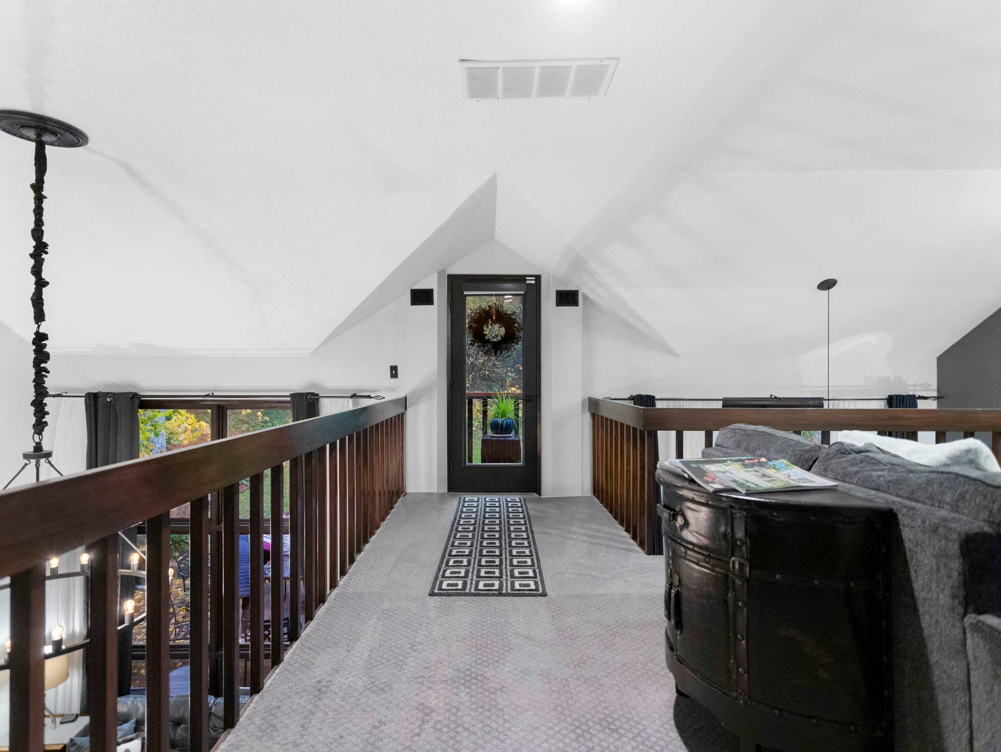 An upper-level loft area with grey carpet and a dark wood railing overlooks the space below, with a view towards a dark-framed door adorned with a wreath. The sloped white ceiling and an air vent are visible.