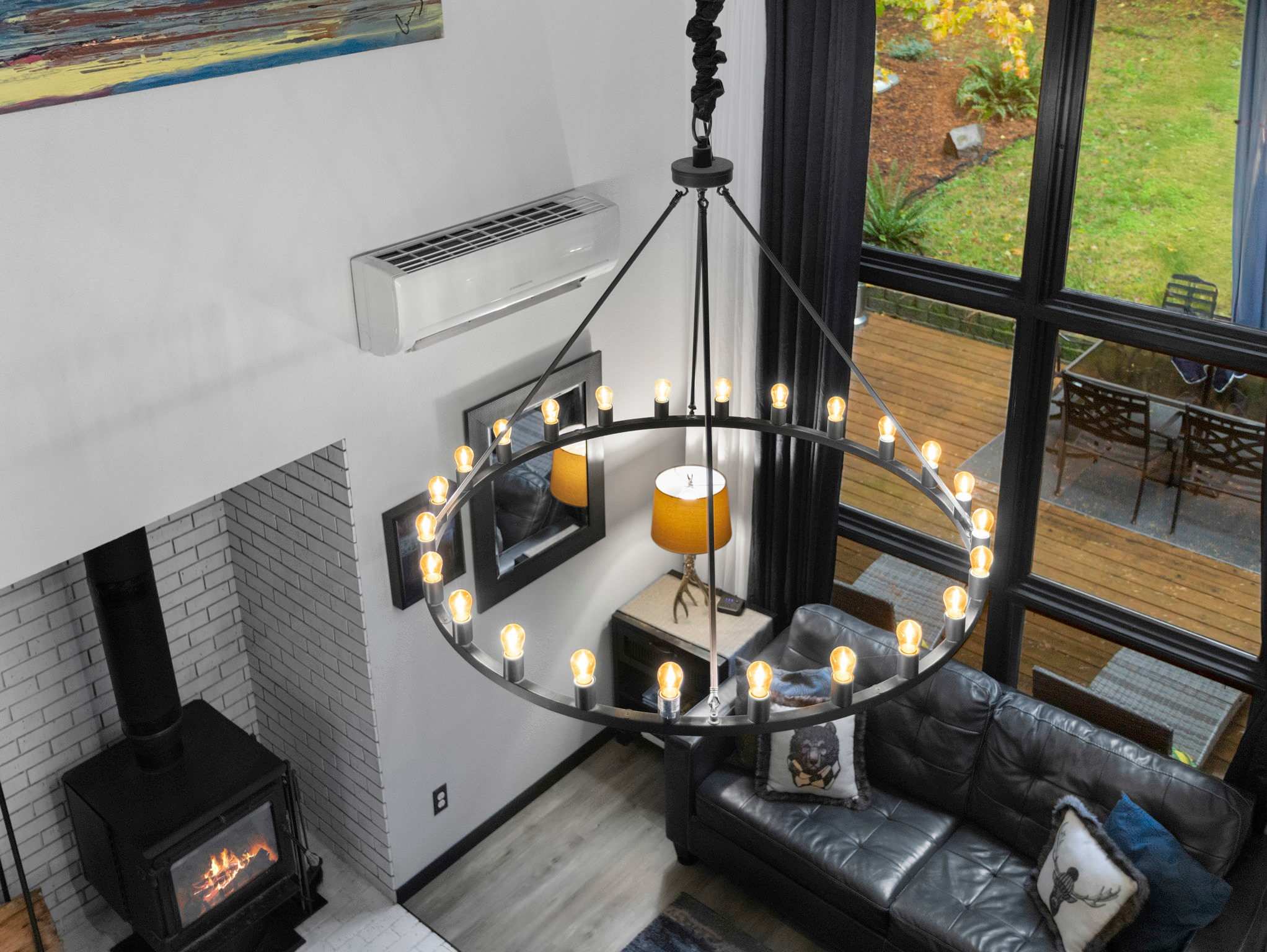 A high-angle view of a living room features a large black leather sofa, a wood-burning stove, and a striking circular chandelier with multiple bulbs. Large windows offer a view of the outdoors, and an air conditioning unit is mounted high on the wall.