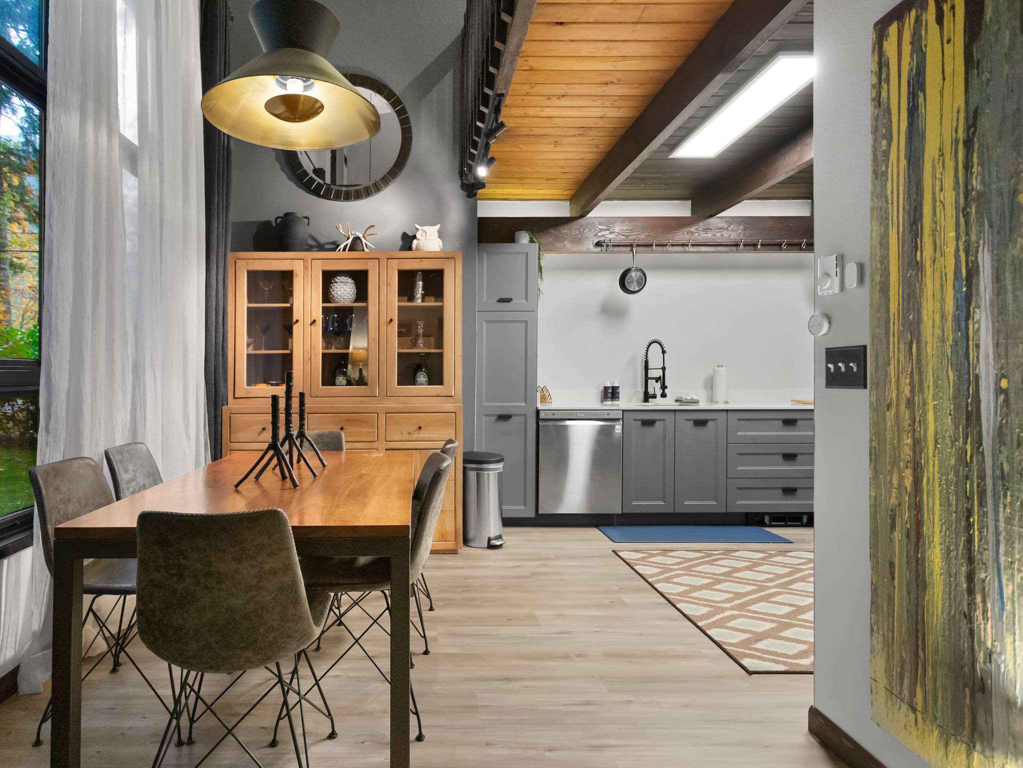 Dining area with a large wooden table and modern chairs, adjacent to a gray kitchen with stainless steel appliances. A wooden hutch displays dishware, and a large round mirror hangs above it.