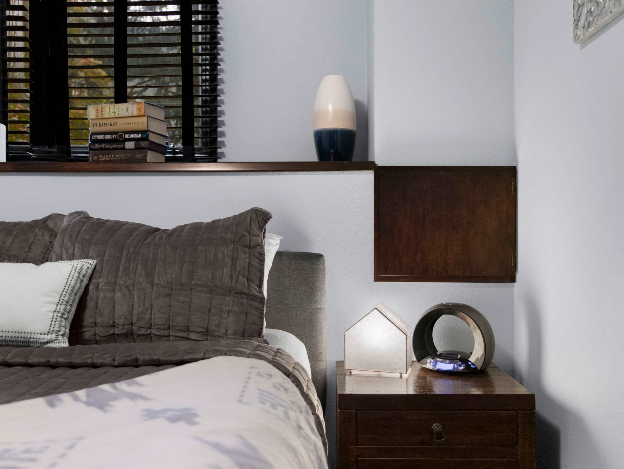 A close-up of a dark wood bedside table in the bedroom, featuring a modern lamp, a small house-shaped decor item, and a two-toned vase on the shelf above.