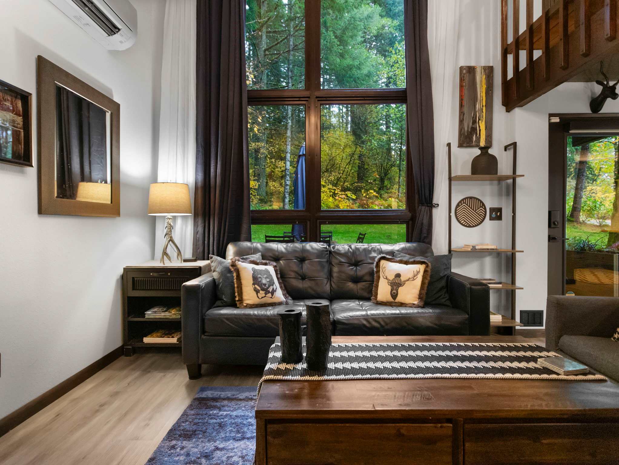 View of the living room with a black leather sofa adorned with decorative pillows, a dark wood coffee table, and a large window showcasing the surrounding forest. Dark curtains frame the window, and a modern lamp sits on a side table.