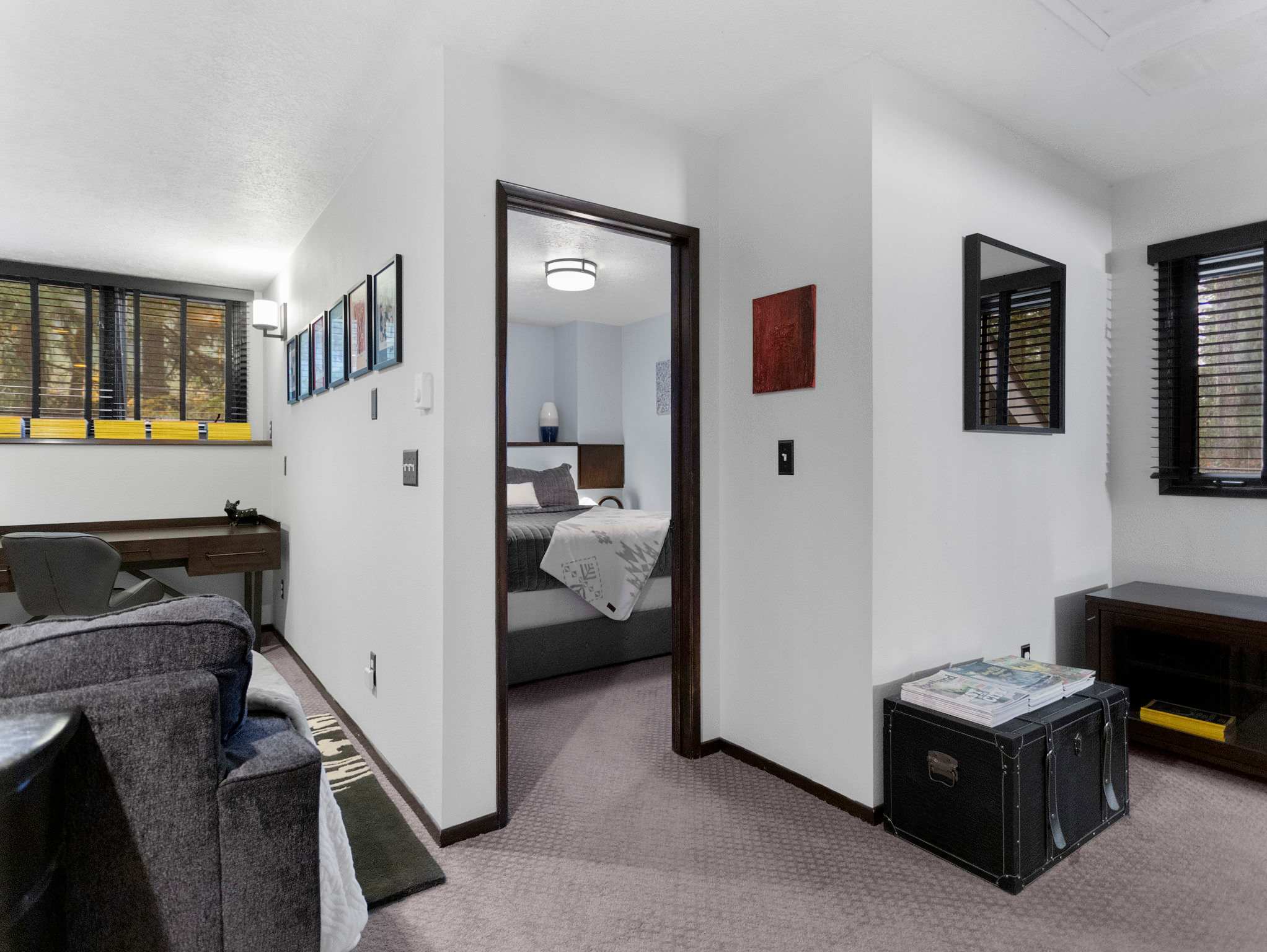 An open loft space features a grey sofa on the left and a glimpse of the desk area in the background. A dark wood doorway leads to an adjacent bedroom, while a black trunk and small table occupy the right side of the frame.