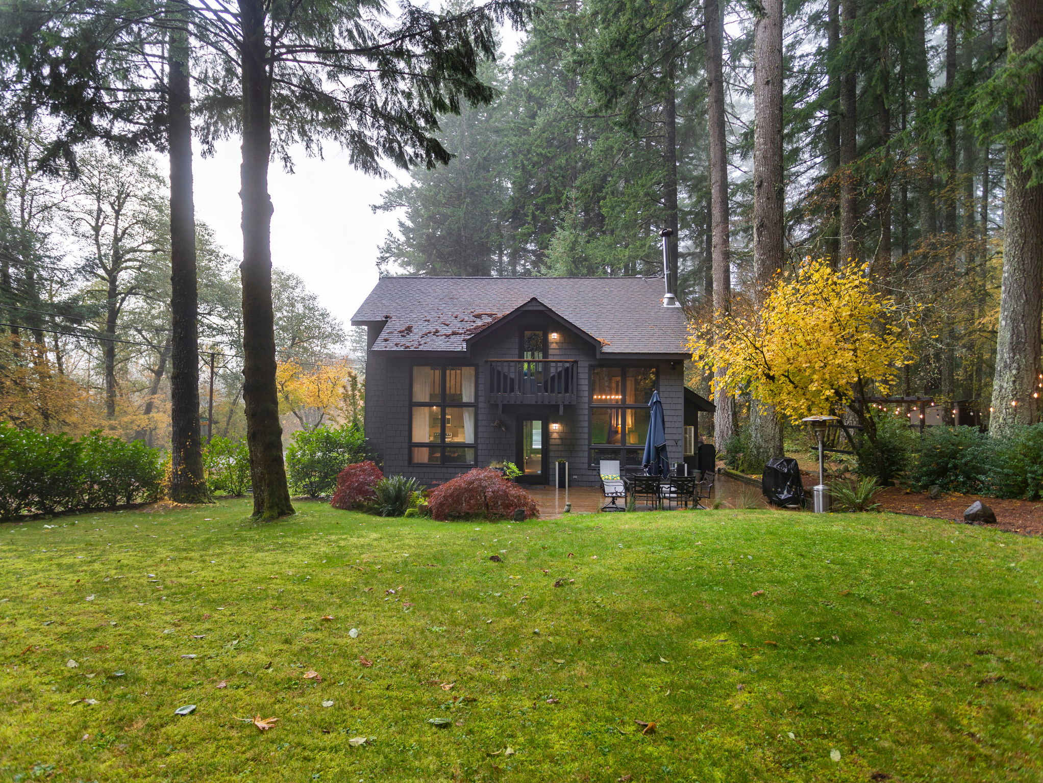 The back of a dark gray house features a small balcony and large windows, overlooking a well-maintained green lawn. Tall evergreen trees and a vibrant yellow-leafed tree frame the property, creating a serene forest setting.