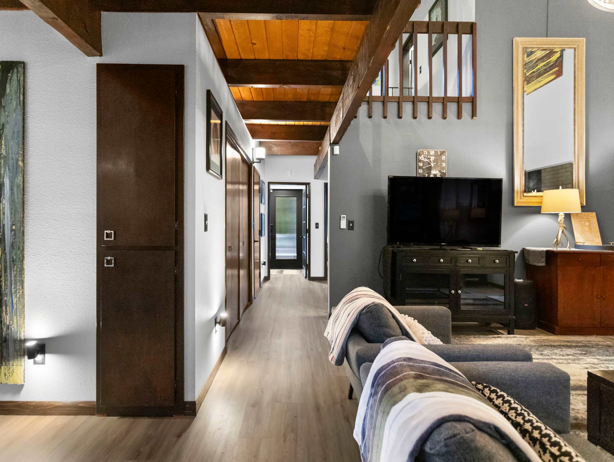 A long hallway with light wood-look flooring and exposed wooden beams leads towards a dark doorway, with a cozy living area visible to the right. The living space features a grey sectional sofa with a striped throw blanket and a dark TV console.