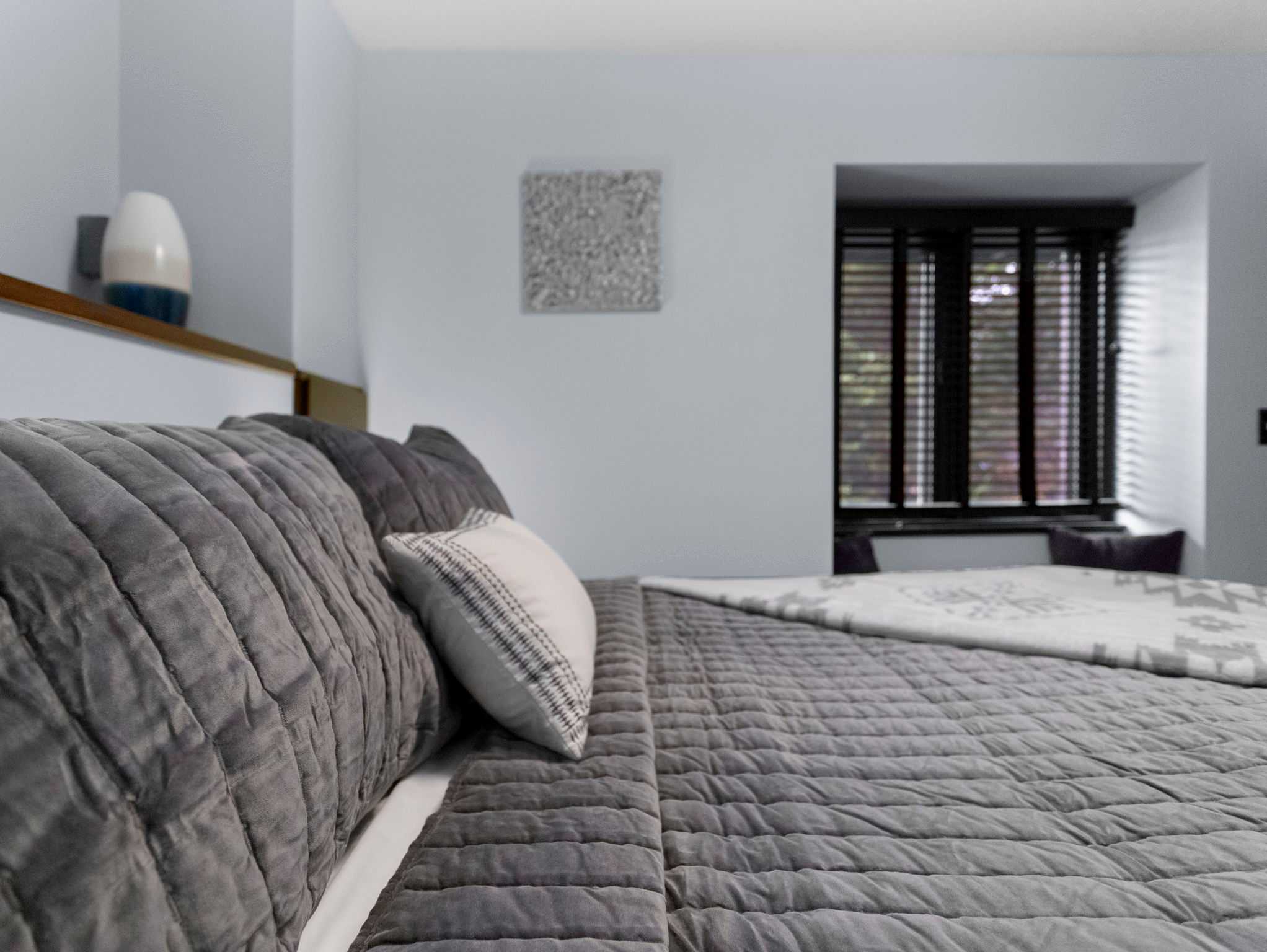 A cozy bedroom featuring a bed with plush grey bedding and a white patterned throw blanket. A window with dark wood blinds allows natural light, and a light blue wall adds a calming touch.
