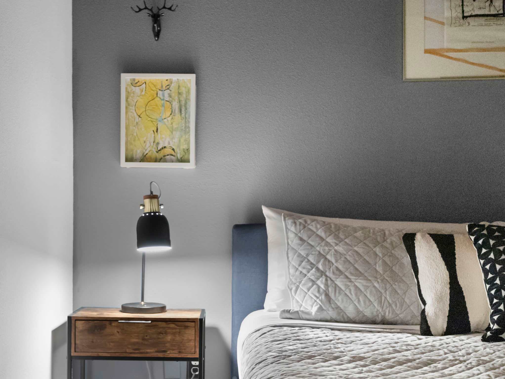 A close-up view of the bedroom shows a dark nightstand with a modern black lamp and a light blue upholstered bed. The grey wall is decorated with a small abstract painting and a deer head wall decor.