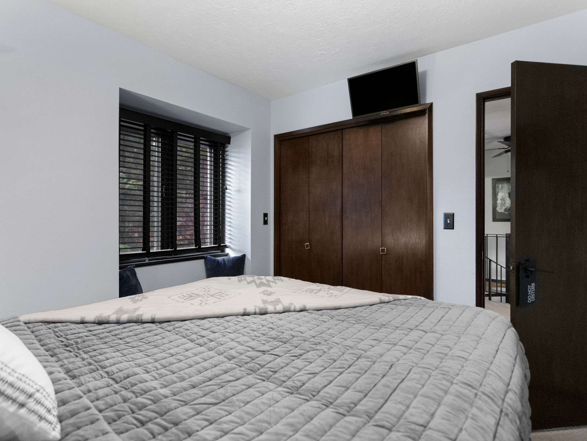 A view of the bedroom showcasing a large dark wood closet with a flat-screen TV mounted above it. The bed with grey bedding and a window with dark blinds are also visible, along with a dark wood door leading out.