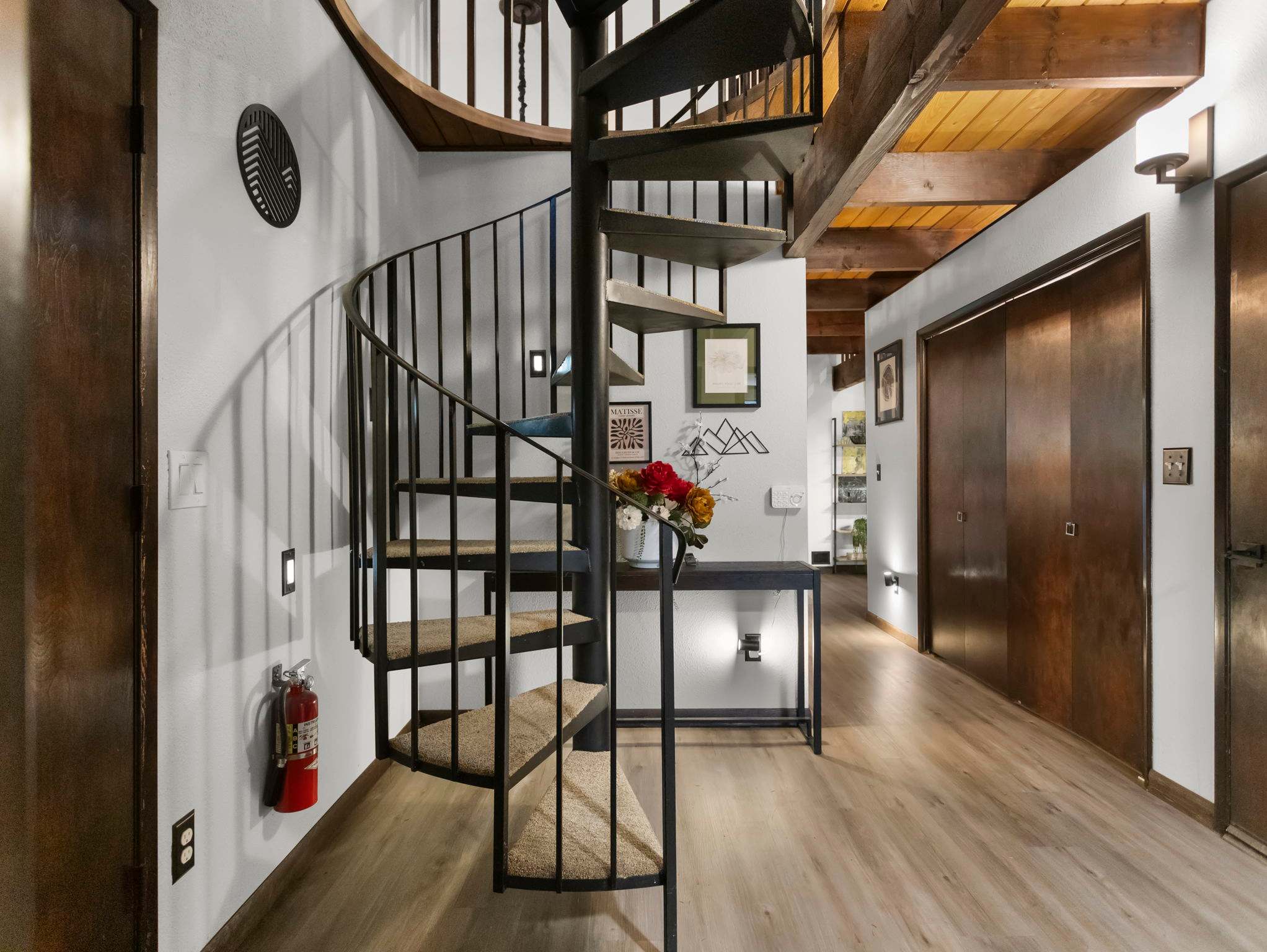 A spacious foyer with light wood flooring and white walls features a black spiral staircase with carpeted treads leading to an upper level. A console table with flowers and framed art decorates the wall.