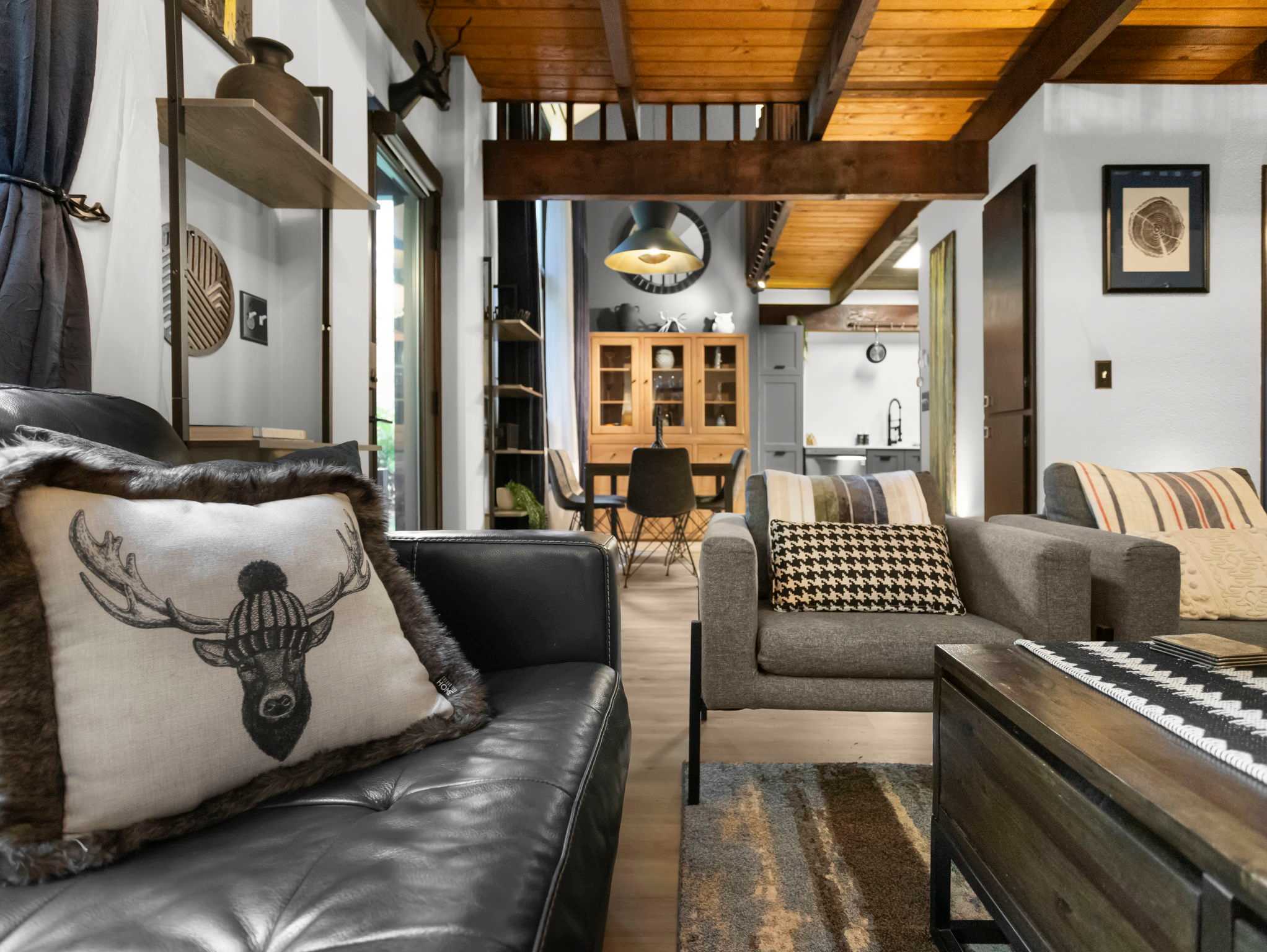 Angled view of the living room with a black leather sofa and gray armchairs, looking towards the dining area and kitchen. Exposed wooden beams and a wooden railing from an upper level add to the chalet aesthetic.