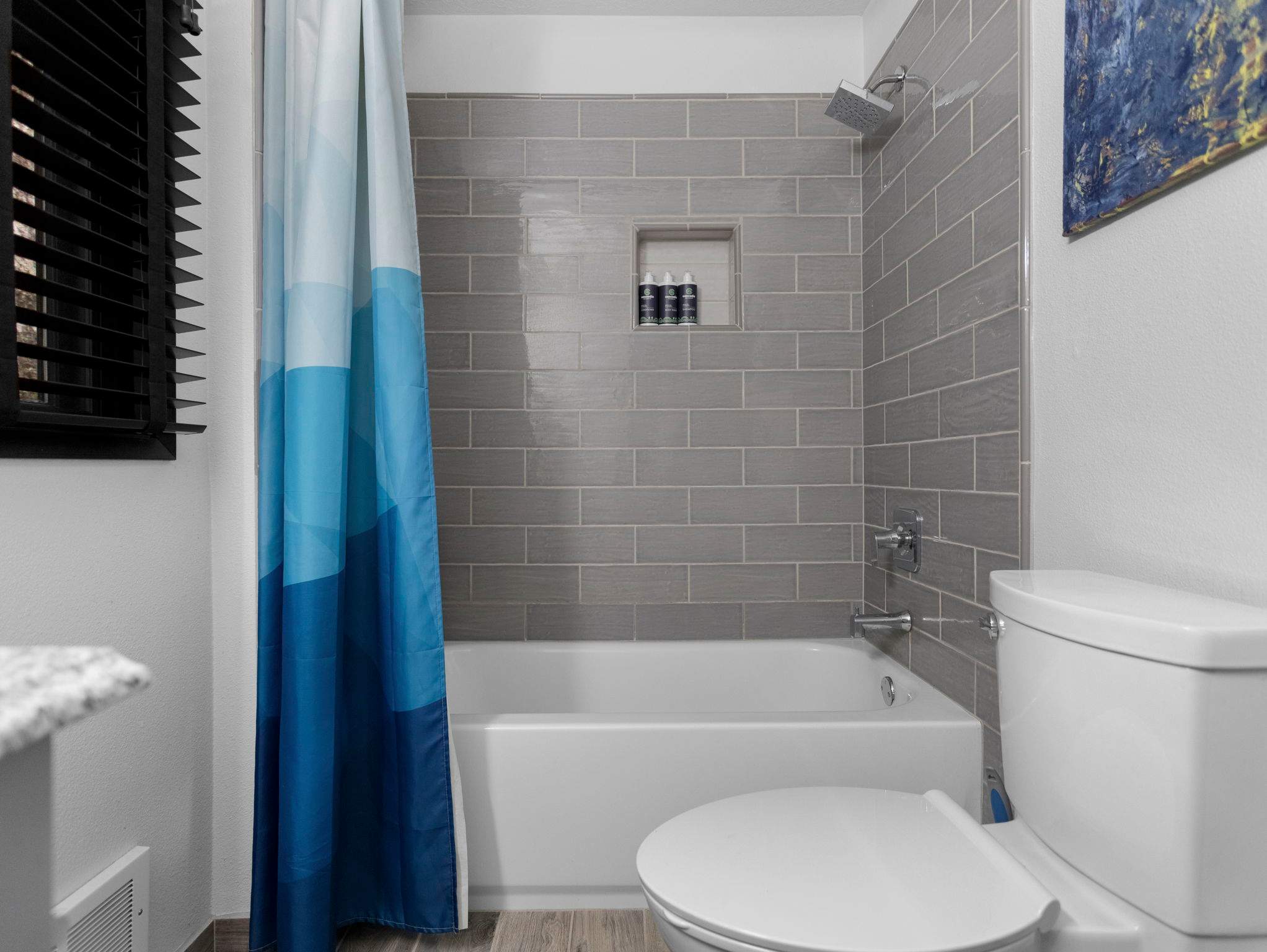 Contemporary bathroom with a white bathtub and shower featuring gray subway tile walls and a built-in niche for toiletries. A white toilet and a blue and white geometric shower curtain are also present.