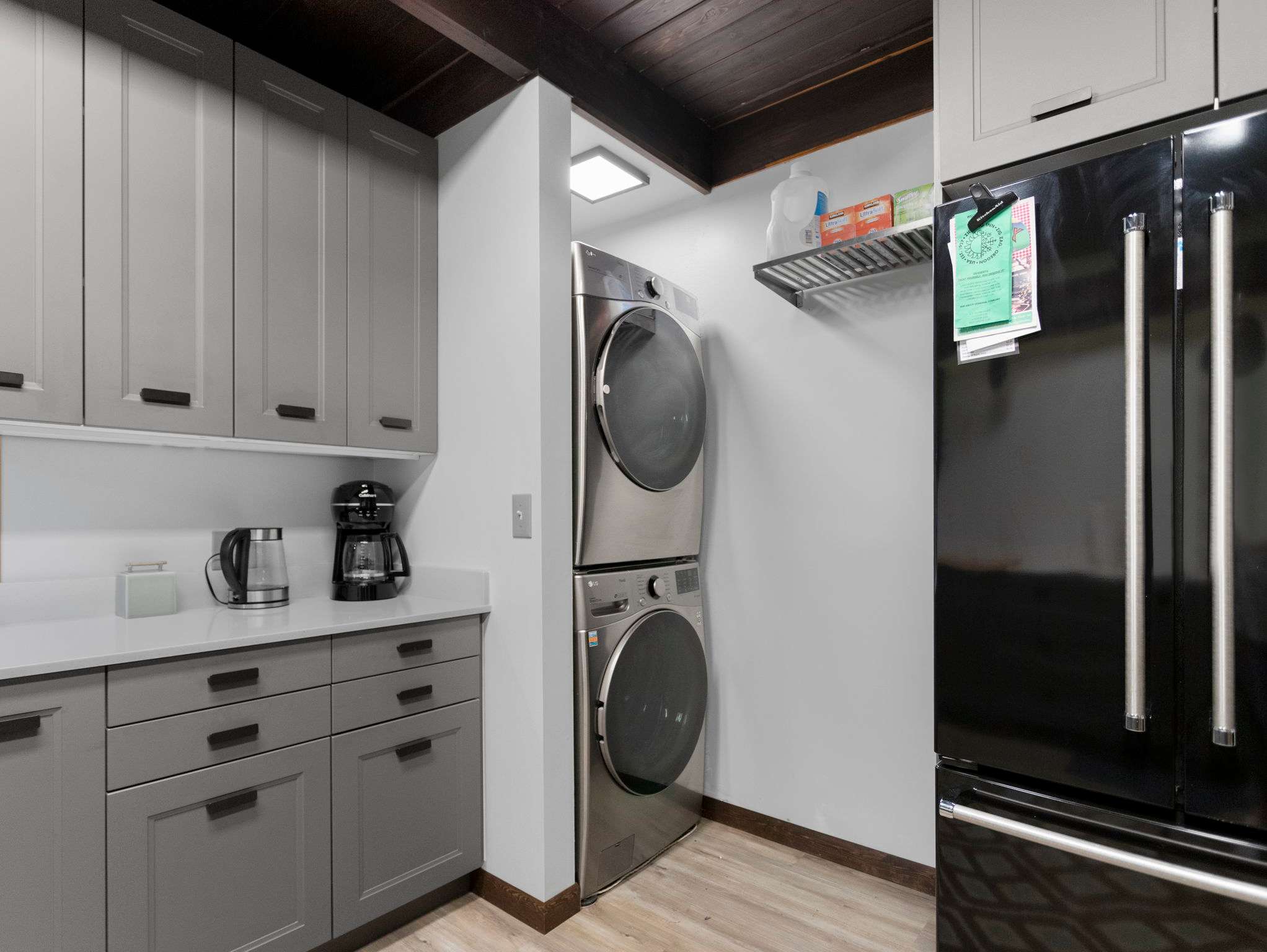 A dedicated laundry nook features a stacked stainless steel washer and dryer unit, neatly tucked beside a black French-door refrigerator. Light grey cabinets and white countertops extend from the adjacent kitchen area, with a wall shelf above the appliances.