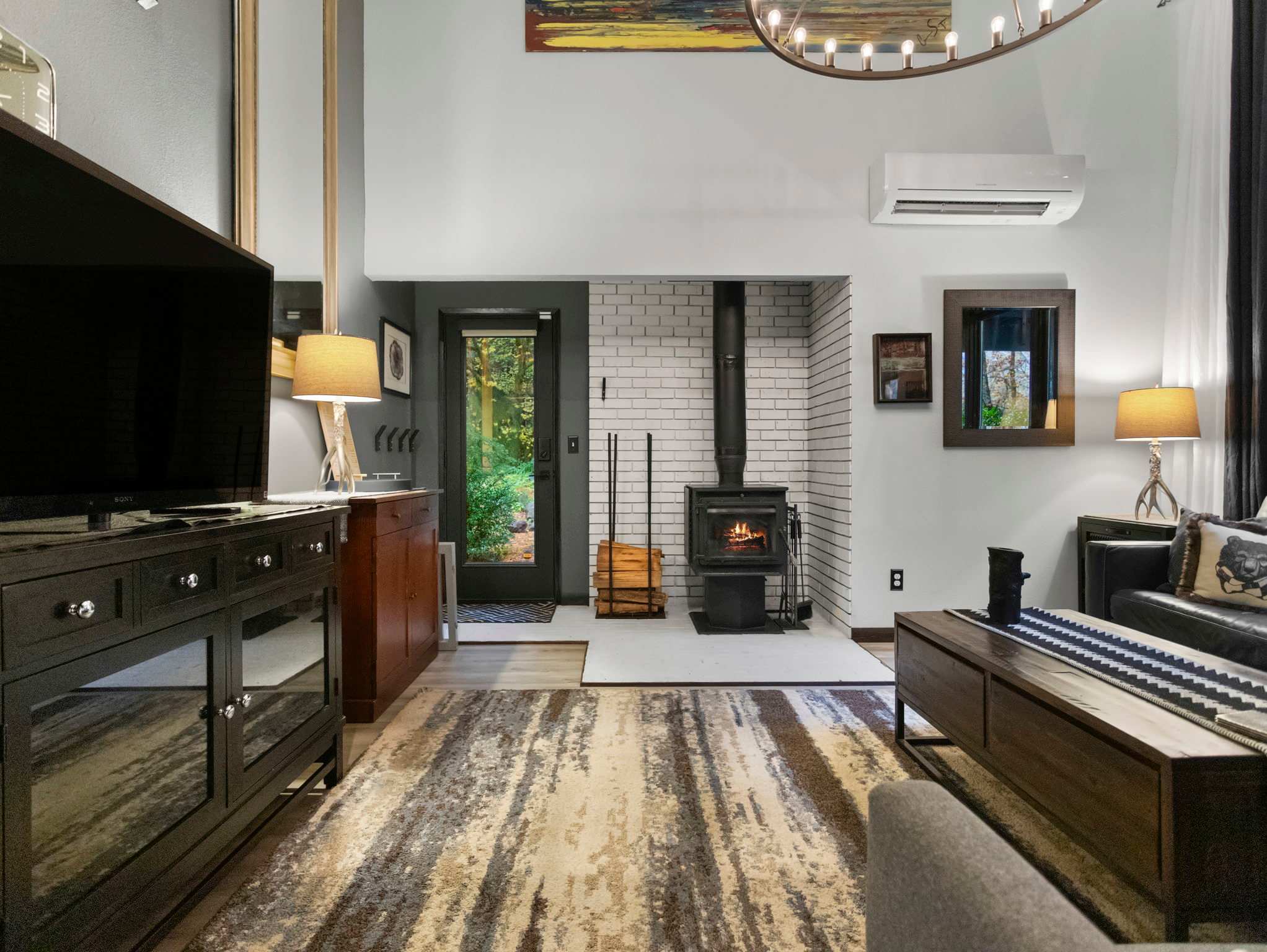 Wide shot of the living room featuring a wood-burning stove with a white brick surround, a large patterned rug, and a dark wood coffee table. A black door leads to the outside, revealing a glimpse of the forest.