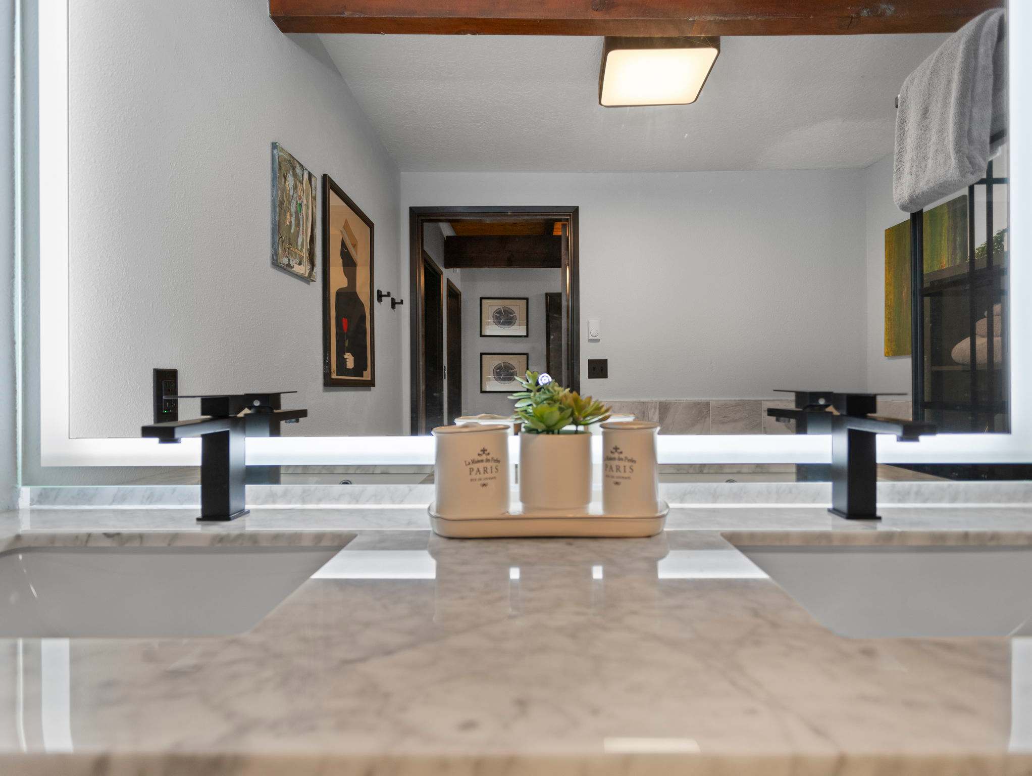 A sleek double vanity in a modern bathroom features a white marble-look countertop with two integrated sinks and black matte faucets. A large illuminated mirror spans the width of the vanity, reflecting the room's dark wood beams and light walls.