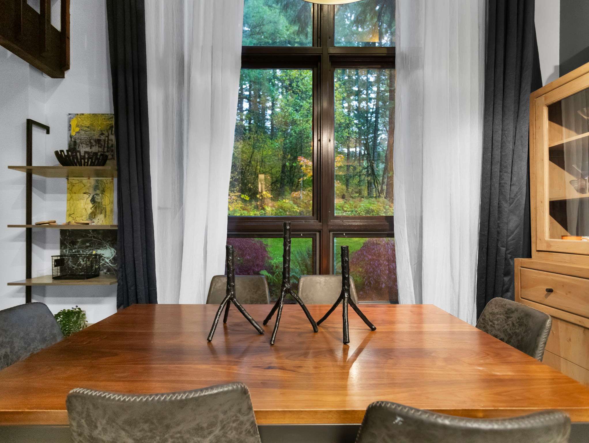 A warm wooden dining table with four grey chairs is centered in front of large windows offering a lush forest view, flanked by white and dark grey curtains. Three black modern candelabras adorn the table, with a dark metal shelving unit visible on the left.