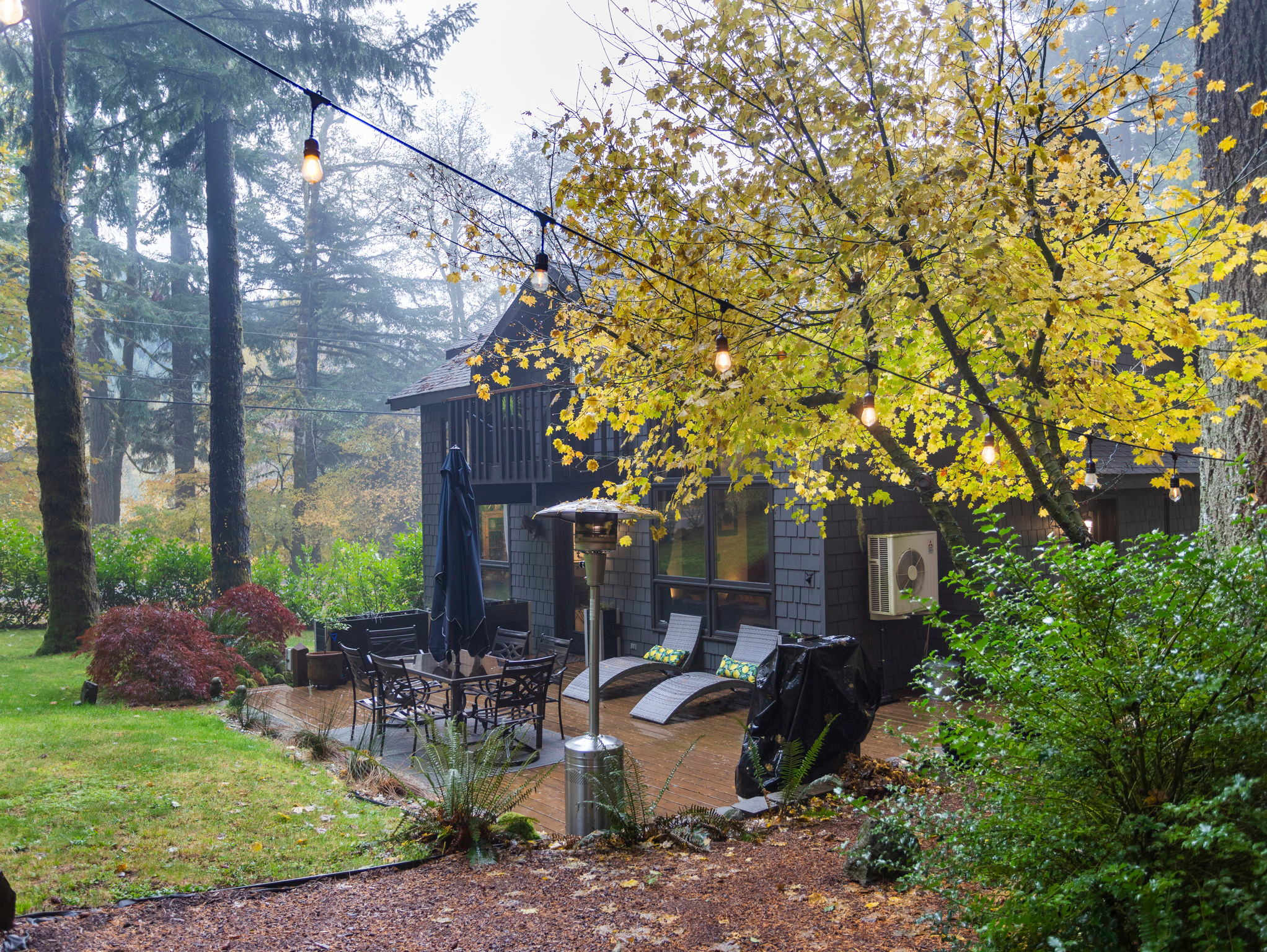 An expansive wooden deck is furnished with an outdoor dining set, lounge chairs, and a patio heater, all illuminated by string lights. The dark gray house with large windows is nestled among tall evergreen and yellow-leafed deciduous trees.