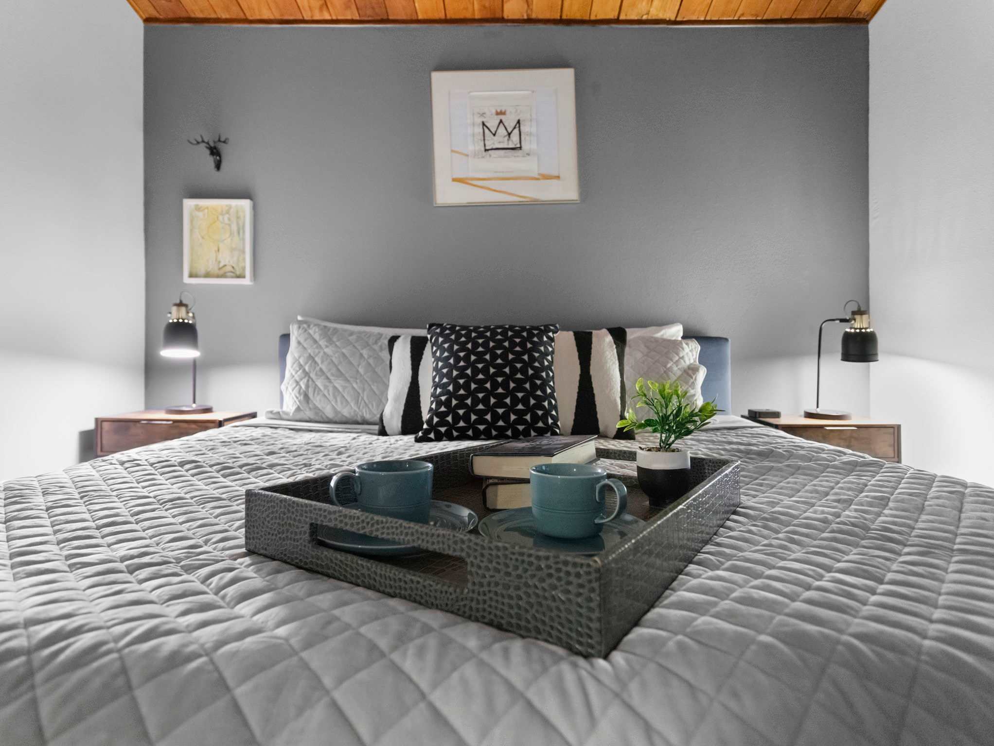 A detailed view of the bed in bedroom-1 features a grey quilted comforter, various pillows, and a decorative tray with two blue cups and a small plant. Exposed wooden beams are visible on the ceiling.