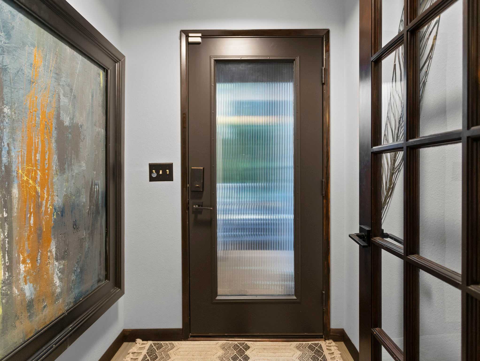 The interior view of the front door, which is dark with a frosted glass panel, and a patterned rug on the floor. A large framed artwork hangs on the wall next to the door.