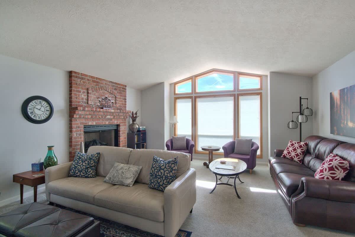 A wide shot of the living room showcasing a brick fireplace, a light beige sofa, a dark brown leather sofa, and two purple armchairs by a large bay window, with a coffee table and a wall clock.