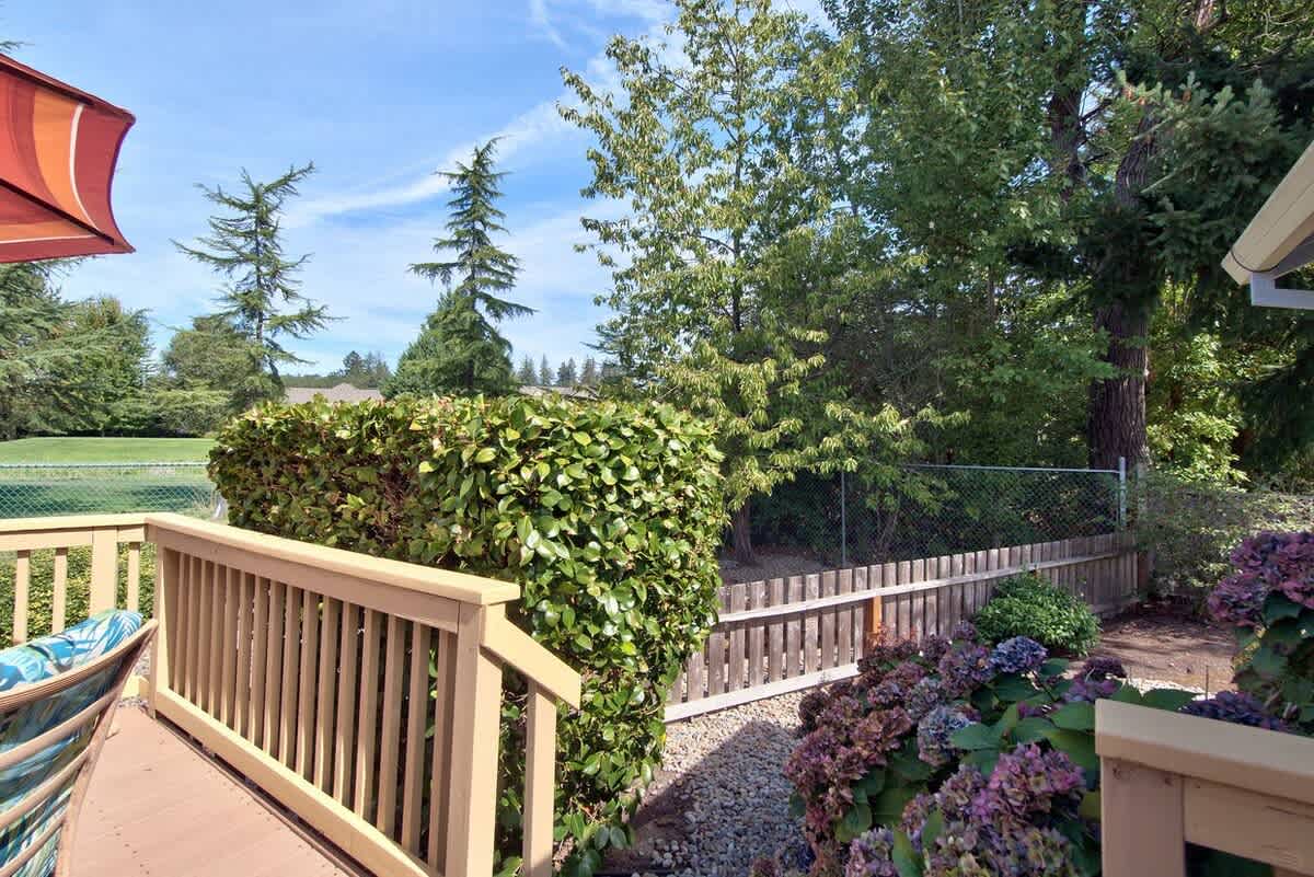 Lush garden area adjacent to the wooden deck, featuring dense green bushes, flowering hydrangeas, and a wooden fence, with tall trees in the background.