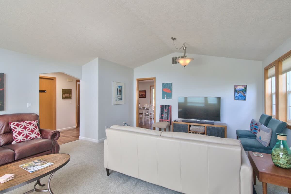 The living room features a light beige sofa facing a large TV on a wooden console, with a dark leather sofa to the left, and an open doorway leading to a dining area visible in the background.
