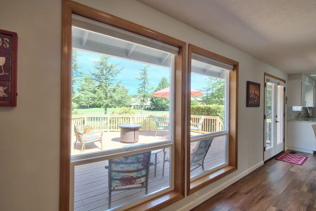 Large windows with light wood trim provide a bright view of the outdoor deck, furnished with seating and an umbrella, and glimpses of the kitchen and dining area inside.