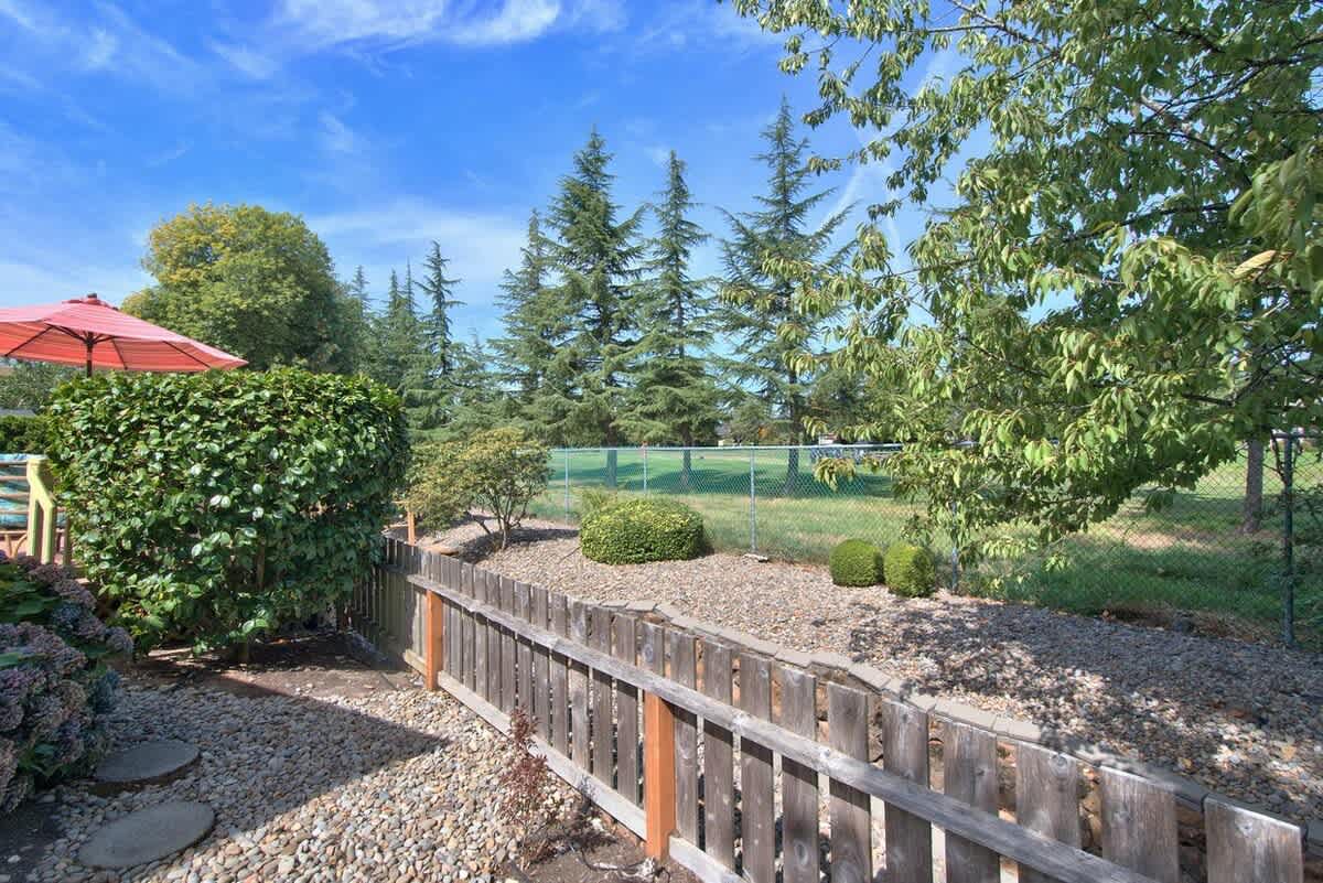 Scenic view of a well-maintained garden with small shrubs and a wooden fence, opening up to a sprawling green lawn and a golf course lined with tall evergreen trees under a bright blue sky.