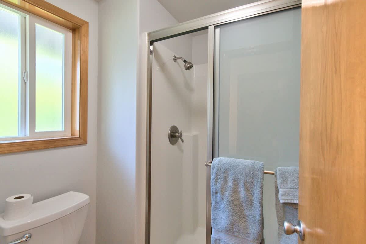 A clean bathroom featuring a white toilet next to a window with wooden trim, and a glass-enclosed shower with a silver showerhead and handle. Gray towels are hanging on a bar inside the shower.