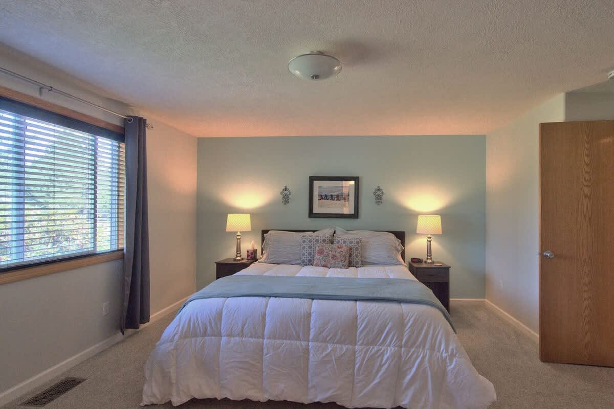 Cozy bedroom with a queen-sized bed featuring white bedding and blue accents, flanked by two nightstands with lamps, and a large window with blinds and curtains.