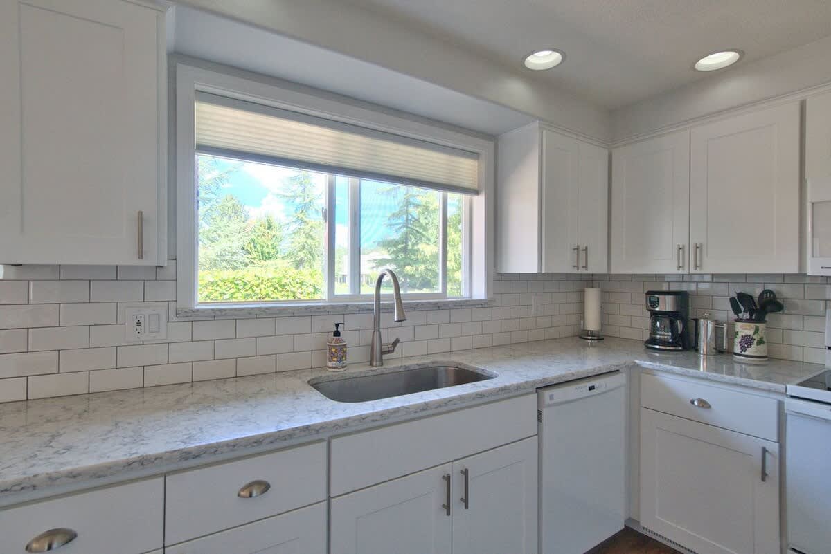 Kitchen sink area with a stainless steel sink, modern faucet, and soap dispenser, set into a light granite countertop with white subway tile backsplash, under a large window with outdoor views.