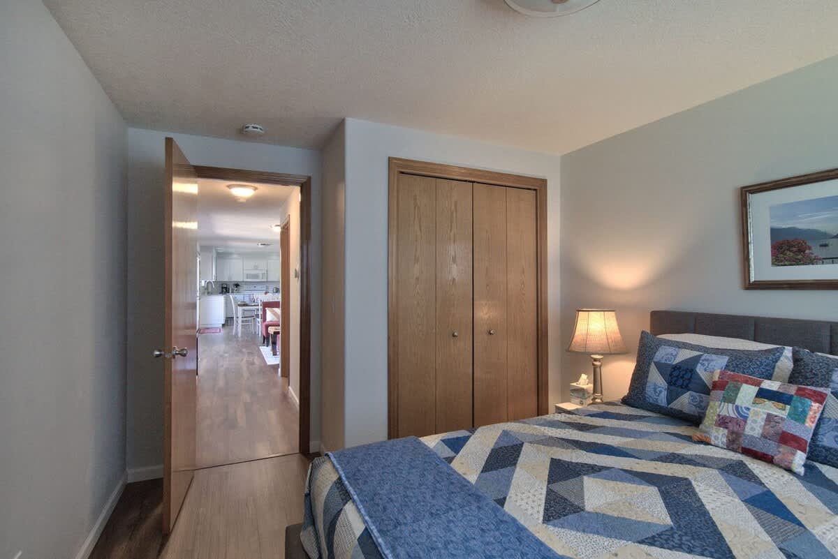Another angle of the bedroom, showcasing a wooden bi-fold closet door and an open doorway leading to a hallway with wood-look flooring. The bed with its blue quilt and a nightstand with a lamp are also visible.