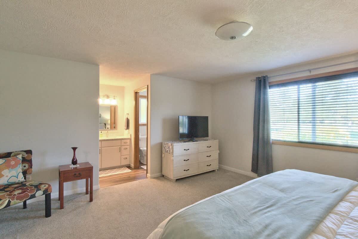 A spacious bedroom with light gray carpet, a large window with blinds and dark curtains, and a white dresser with a TV. An open doorway leads to an en-suite bathroom with a vanity.