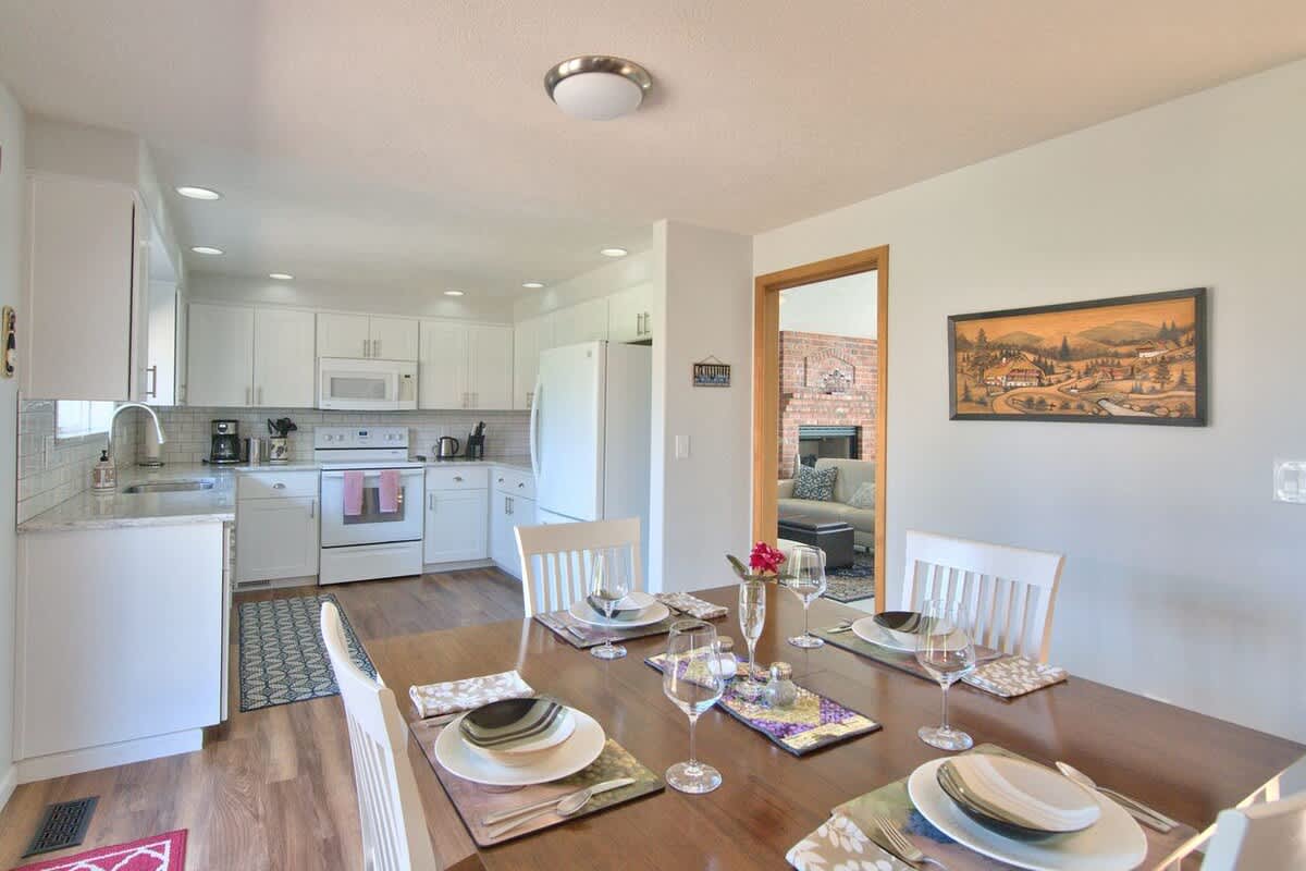 Dining area with a dark wood table and white chairs, set with place settings, looking towards a modern white kitchen with recessed lighting and a doorway to another room.