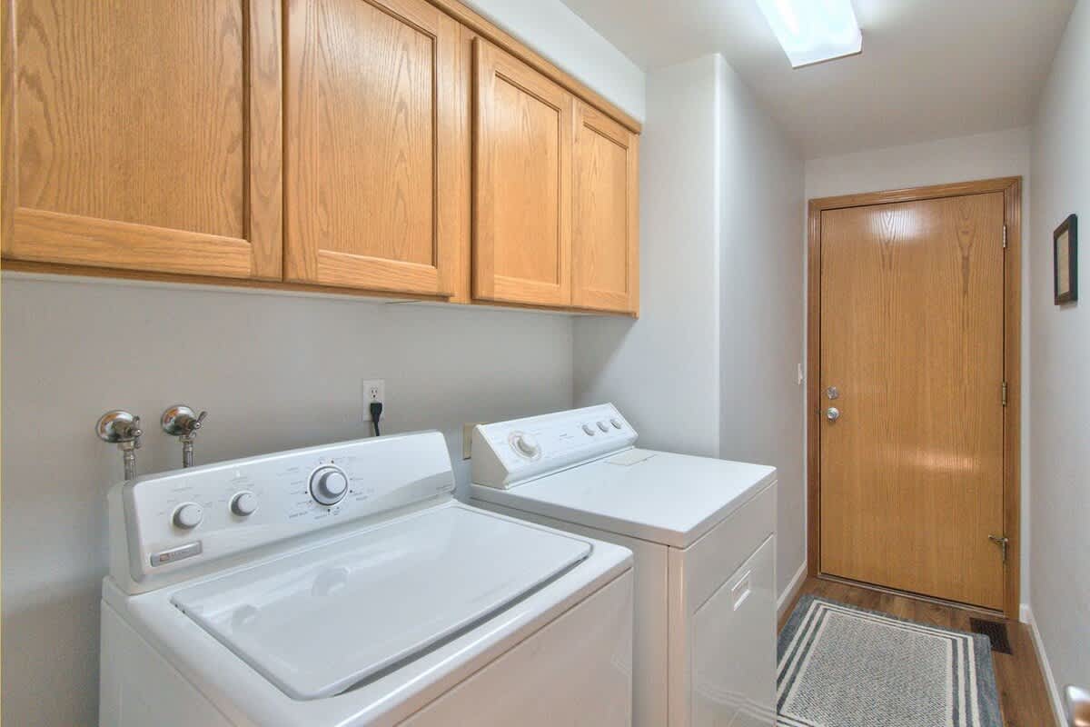 A functional laundry room equipped with a white top-loading washing machine and a matching dryer, set beneath light wood cabinets. A wooden door leads to another area of the house.