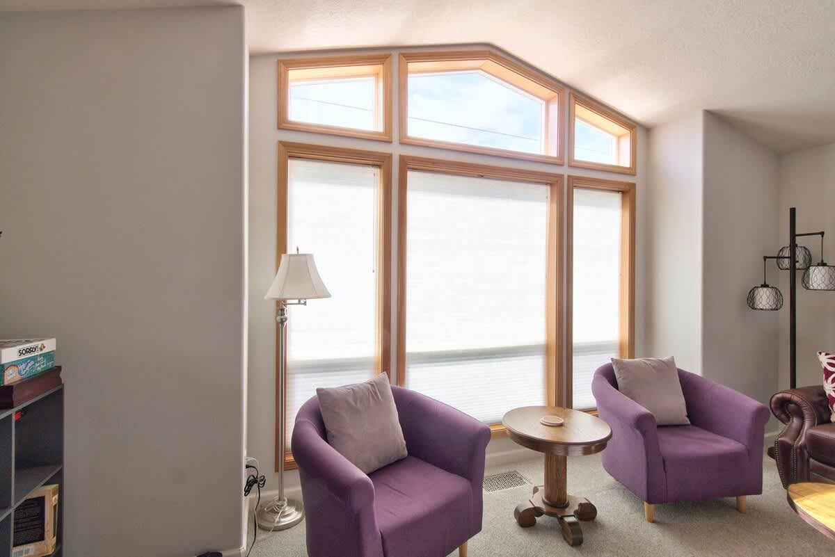 A close-up view of the living room's large bay window with wooden frames and white blinds, featuring two purple armchairs with throw pillows and a small round wooden side table between them.