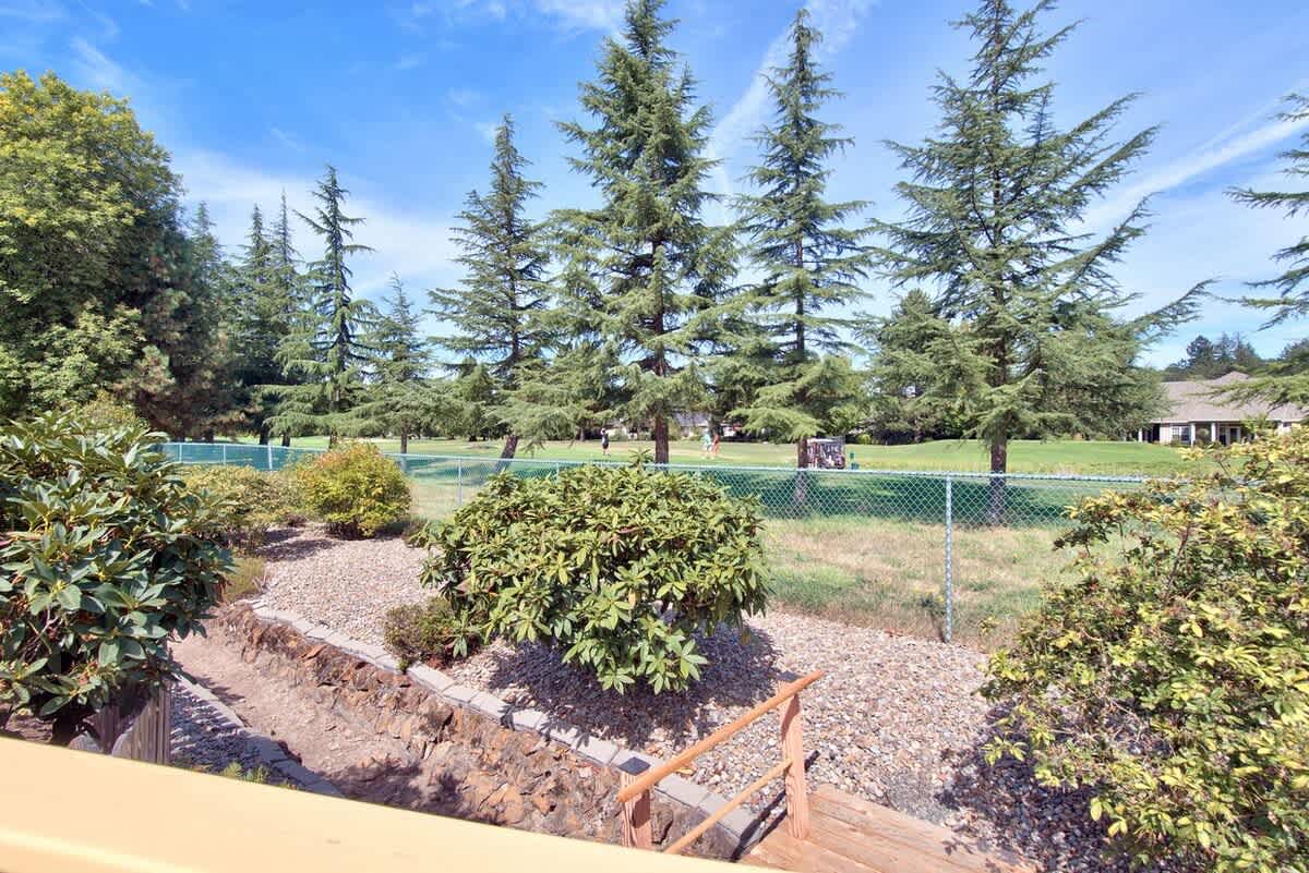 View from the deck showing a landscaped garden area with rocks and shrubs, leading to a chain-link fence bordering a vast green golf course with tall evergreen trees.