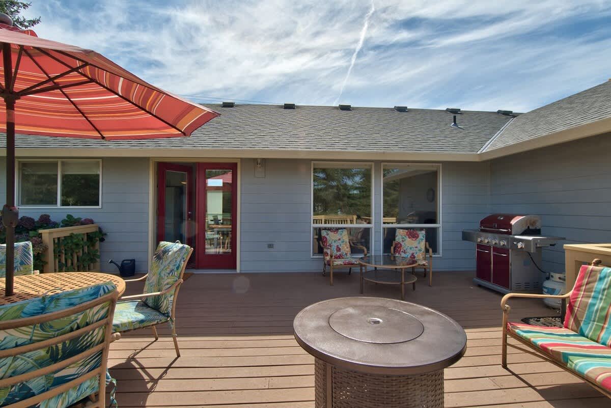 Expansive wooden deck with a red gas grill, a fire pit table, and various outdoor seating, set against the light gray siding of the house with large windows and a red door.