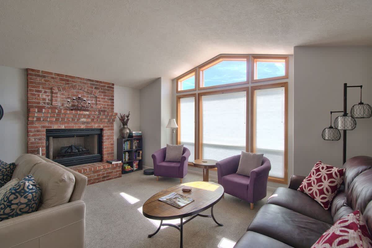 A spacious living room with a brick fireplace, a light beige sofa, and a dark brown leather sofa, centered around a large bay window with wooden frames and white blinds, flanked by two purple armchairs.