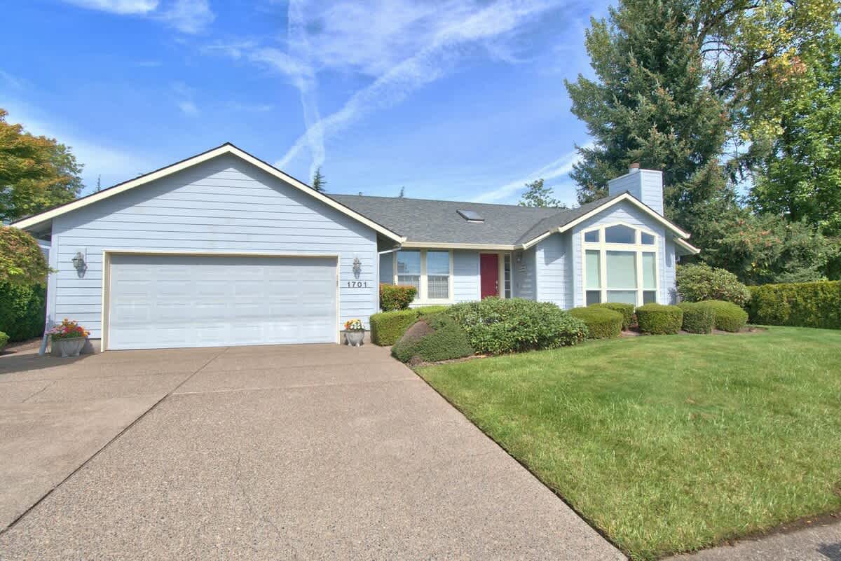 Single-story light blue house with a gray garage door, a red front door, and well-maintained green lawn and landscaping under a bright blue sky.