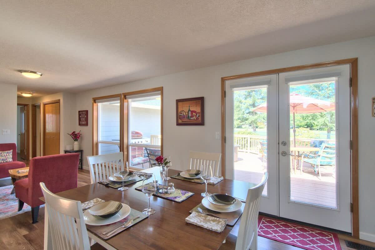 Bright dining area with a dark wood table set for four, surrounded by white chairs, featuring large windows and glass doors leading to an outdoor deck.