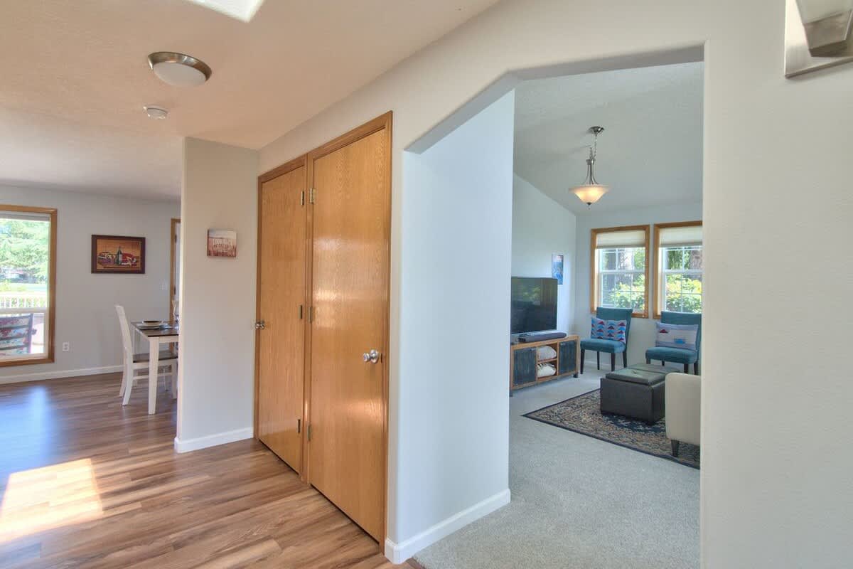 A bright entryway with light wood laminate flooring, a double-door closet, and an arched doorway leading to a carpeted living room with vaulted ceilings and large windows.